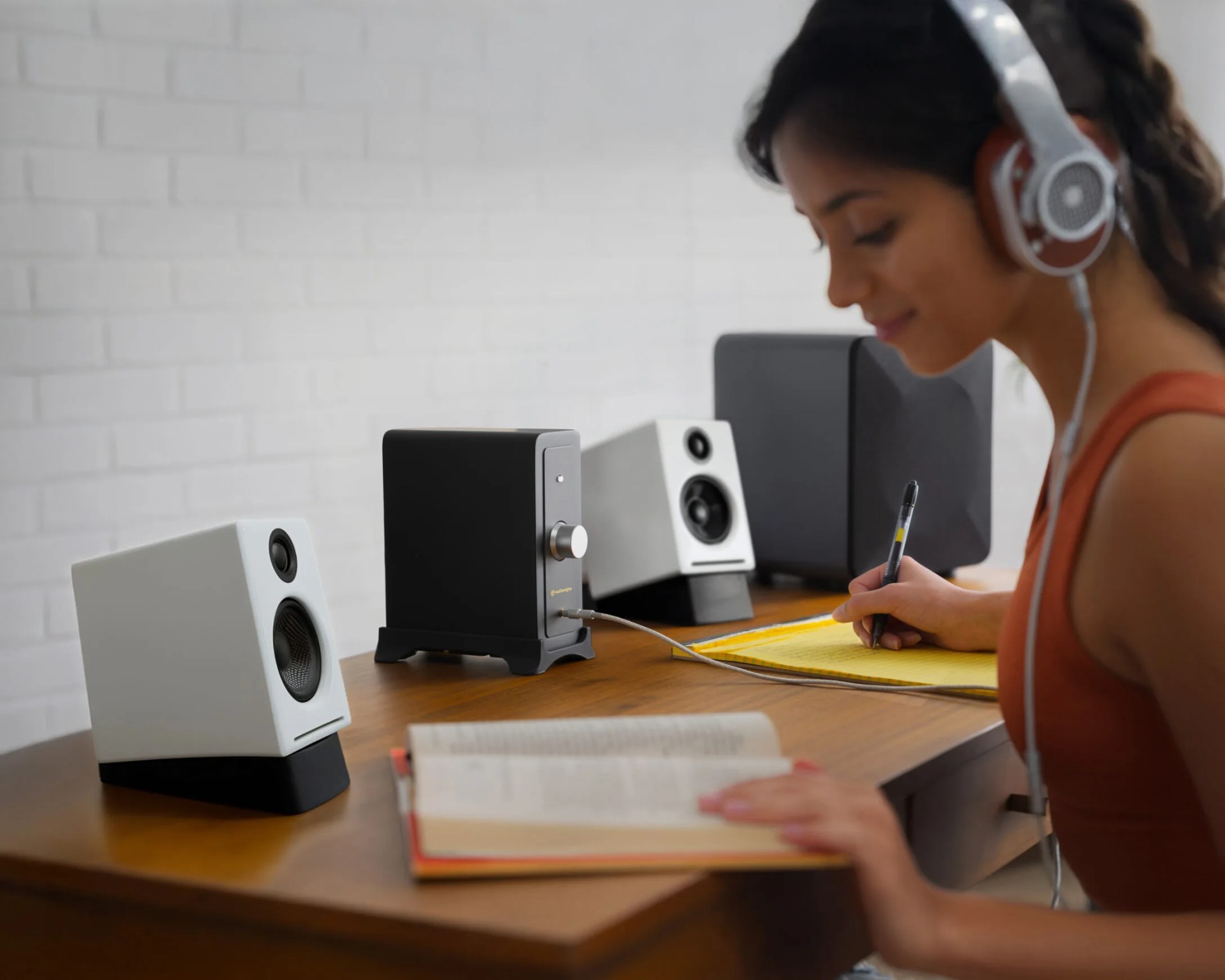 A young woman wearing large over-ear headphones is sitting at a wooden desk, writing on a yellow notepad with a pen. On the desk are two white speakers with black bases, a black audio amplifier with a silver knob, and a black subwoofer. An open book is also on the desk in front of her. The background features a white brick wall.