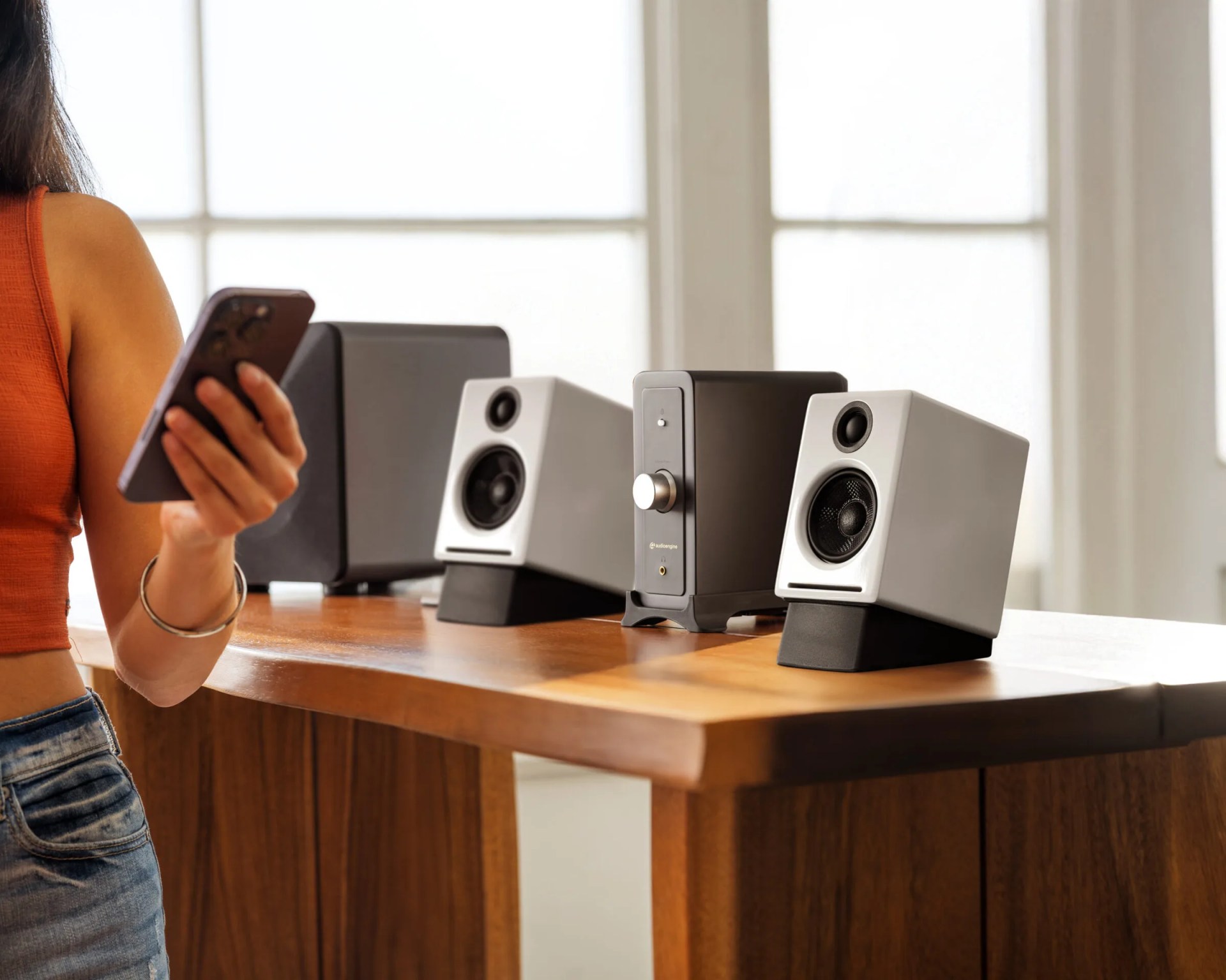 A wooden table with a set of modern audio speakers and a subwoofer, featuring a black and white color scheme. A person wearing a sleeveless orange top and blue jeans is holding a smartphone near the table. Bright natural light comes through large windows in the background.