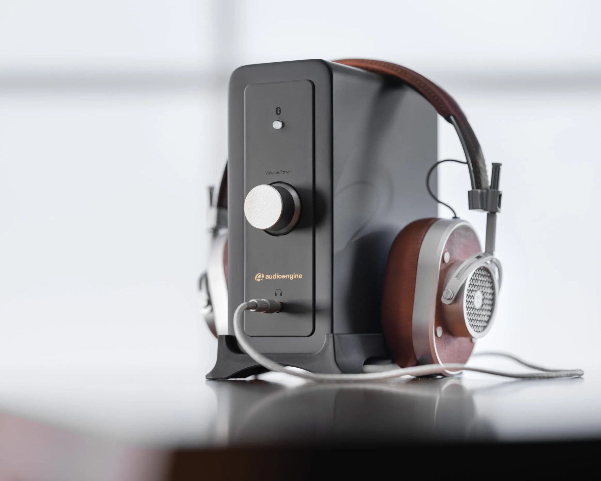 A black Audioengine amplifier with a large silver volume/power knob on the front, connected to a pair of over-ear headphones with brown ear cups, silver metal accents, and a brown leather headband. The headphones are resting on the amplifier, which is placed on a reflective surface.