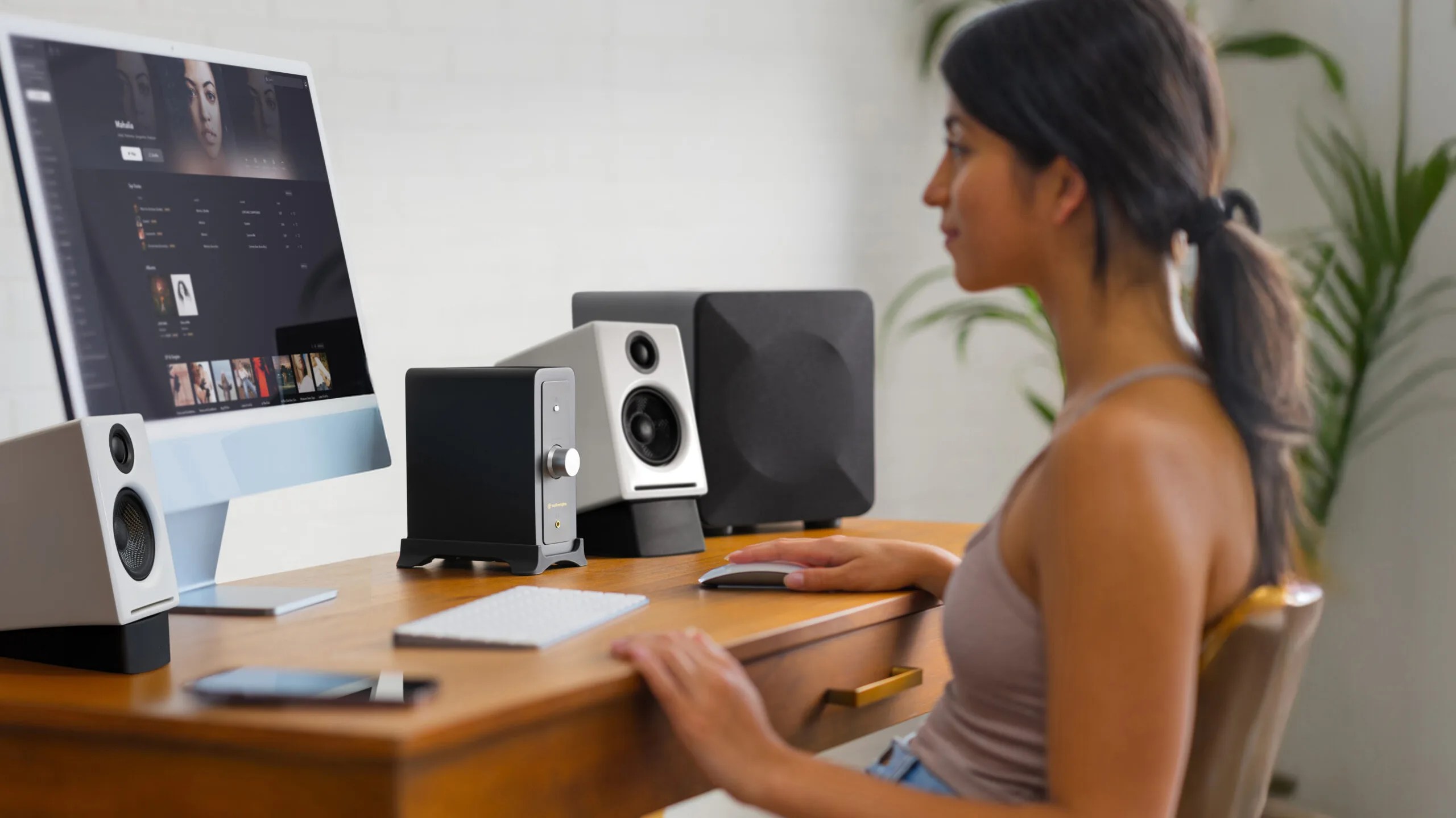 A woman with dark hair tied in a ponytail sits at a wooden desk using a computer with a large monitor displaying a music or media application. On the desk are white speakers, a black audio amplifier, a keyboard, a mouse, and a smartphone. There are green plants in the background.