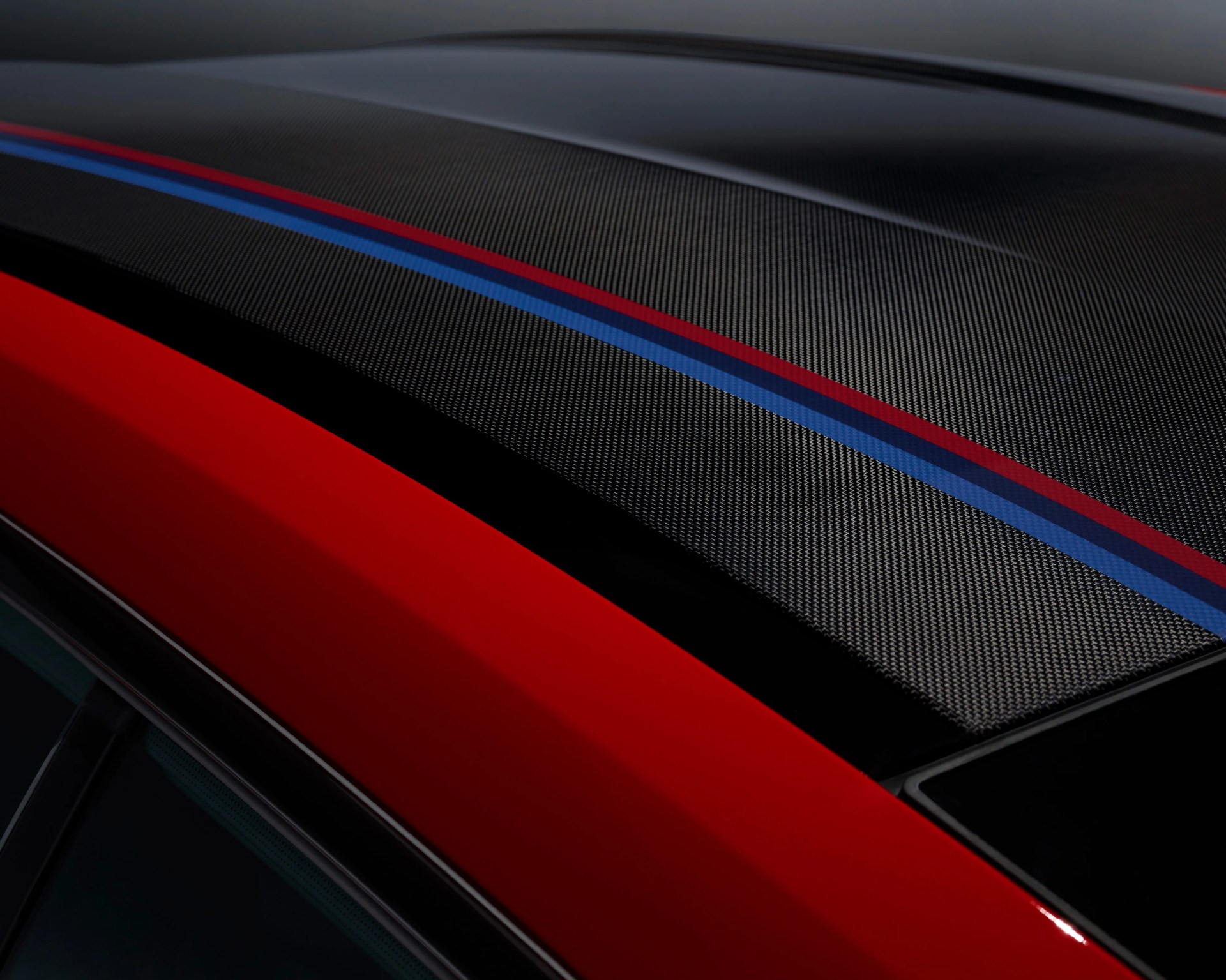 Close-up of a car roof featuring a carbon fiber texture with two racing stripes, one red and one blue, running parallel along the center. The car body visible below the roof is painted in a bright red color.