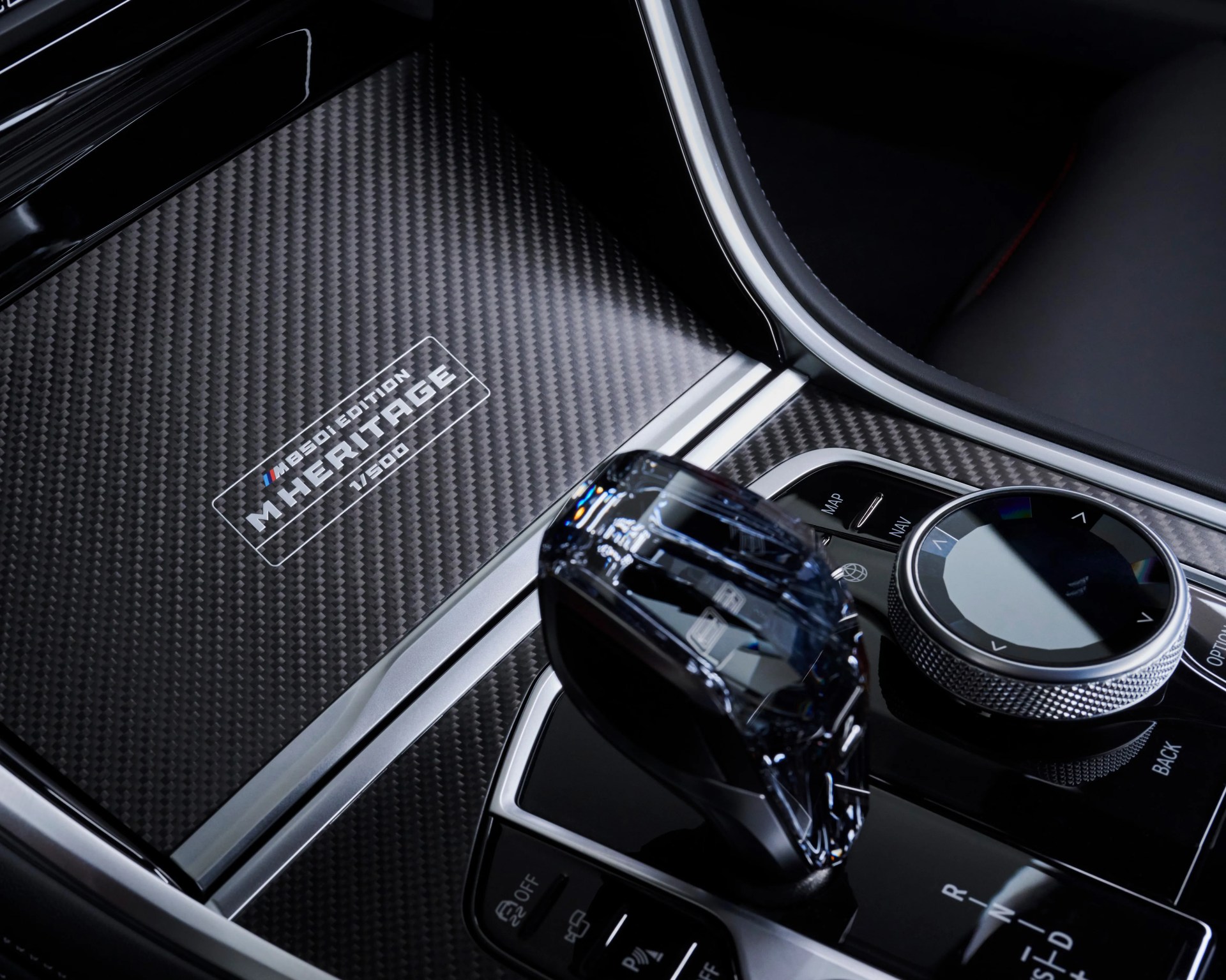 Carbon fiber center console panel with "M850i Edition M Heritage 1/500" badge, crystal gear shifter, and control dial with buttons for map, navigation, options, and back in a luxury car interior.
