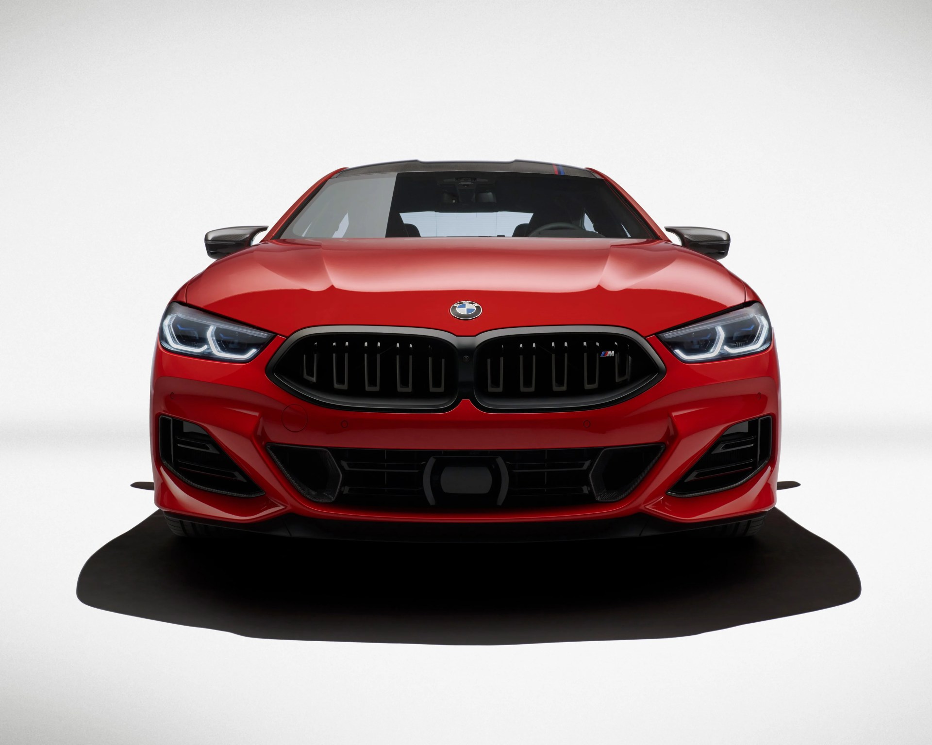 Front view of a red BMW M series car with illuminated LED headlights, a black kidney grille featuring the M badge, and aggressive air intakes on the bumper, set against a plain white background.