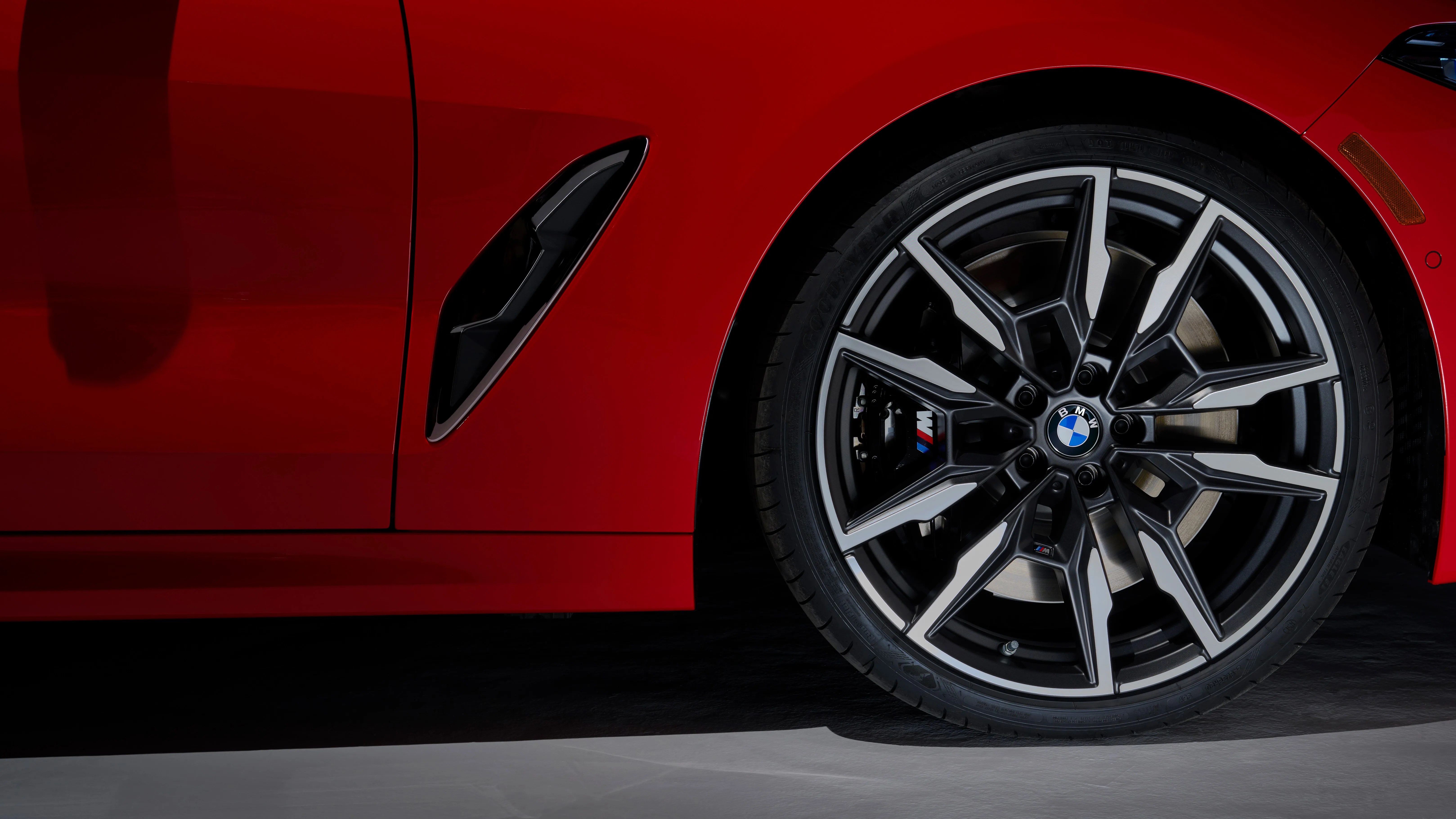 Close-up of a red BMW car's front wheel and part of the side panel, featuring a black and silver multi-spoke alloy rim with the BMW logo in the center and an M badge on the brake caliper. The side panel includes a black air vent detail.