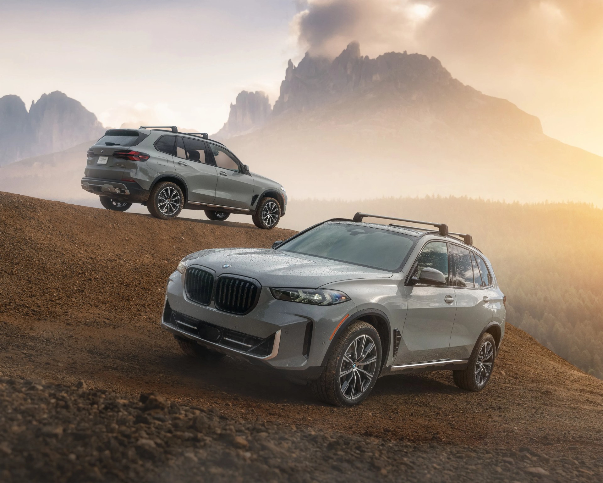 Two silver BMW SUVs are positioned on a dirt hill with a mountainous landscape in the background. The SUV in the foreground faces slightly to the right, showing its front and side, while the SUV in the background is angled to the left, showing its rear and side. Both vehicles have roof racks and sporty alloy wheels. The scene is set during a misty, golden hour with soft sunlight illuminating the area.