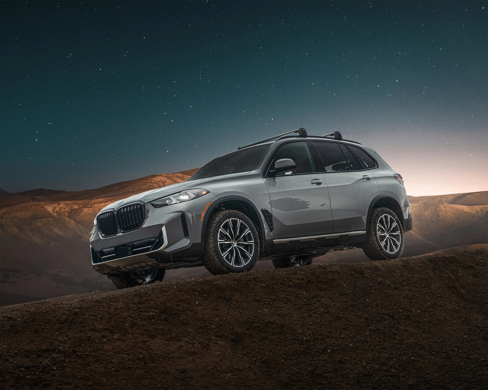 Silver BMW SUV parked on a dirt hill with a starry night sky and mountains in the background. The vehicle has black roof rails and sporty alloy wheels.