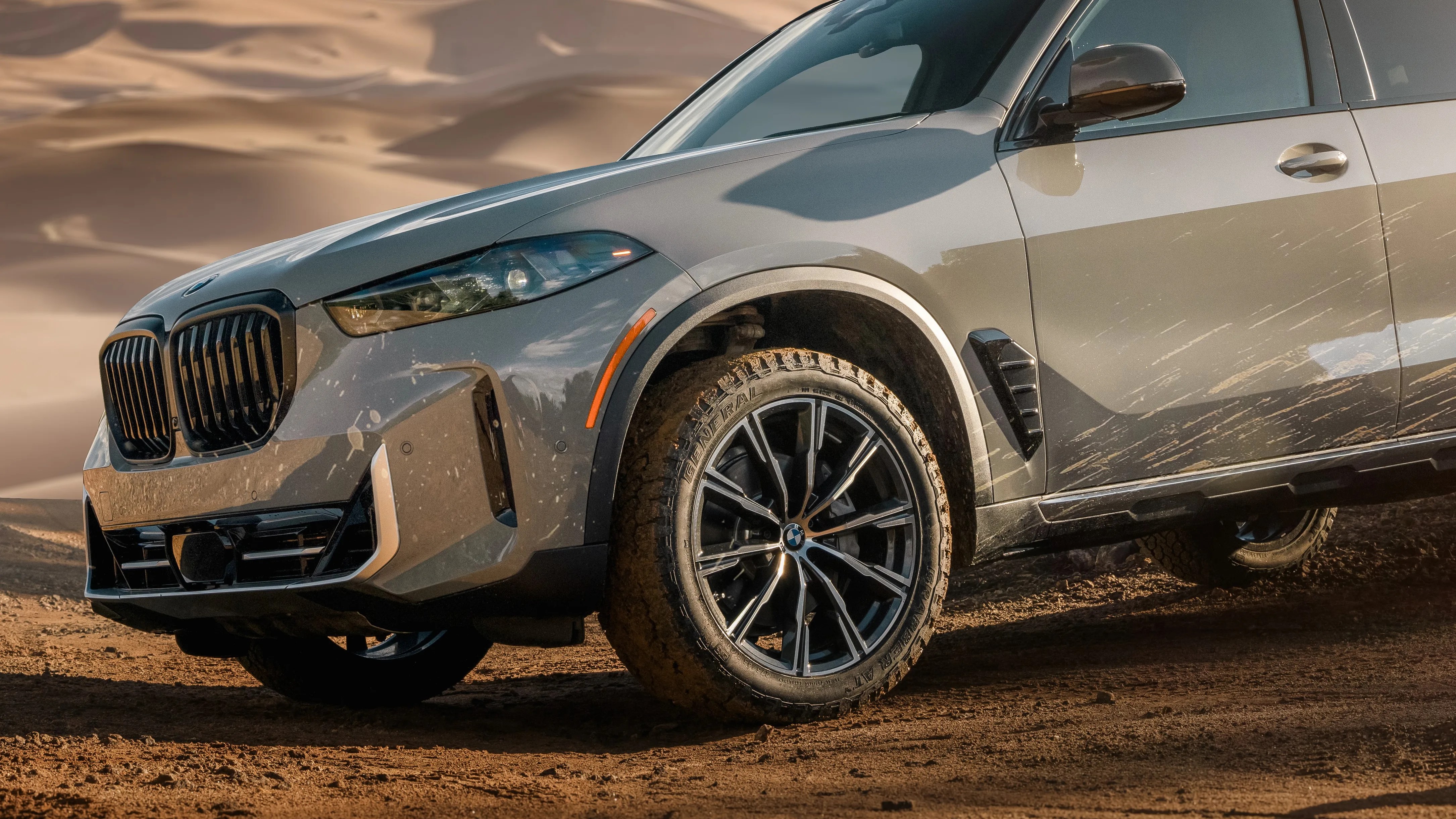 Front side view of a gray BMW SUV with mud splatters on the body and tires, parked on a dirt surface with sand dunes in the background. The vehicle features black alloy wheels and a distinctive kidney grille.