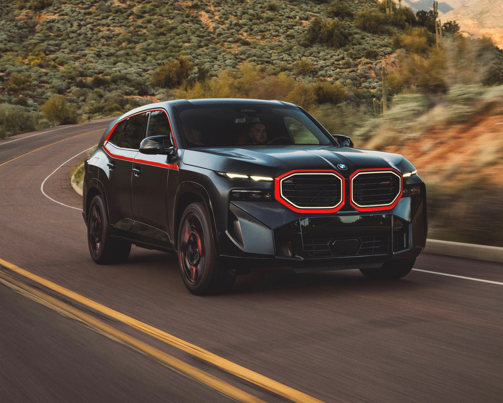 Black BMW SUV driving on a winding road with desert vegetation in the background. The vehicle features distinctive red accents outlining the kidney grille and along the side windows, with sleek, narrow headlights and sporty black wheels.