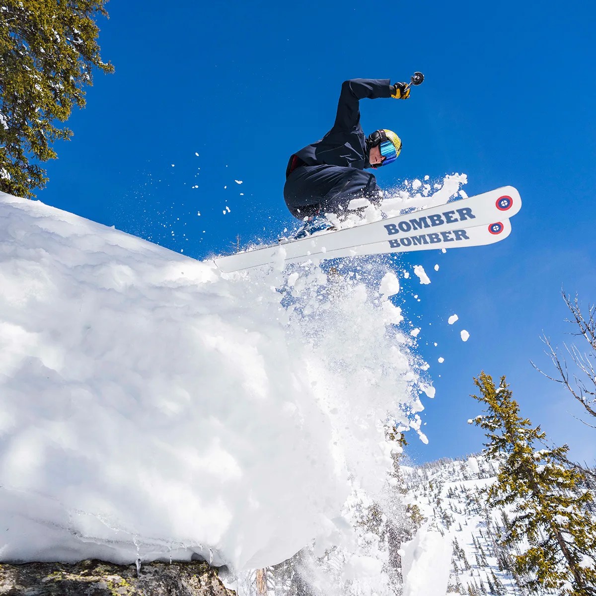 man jumping off snowy hill wearing bomber skis