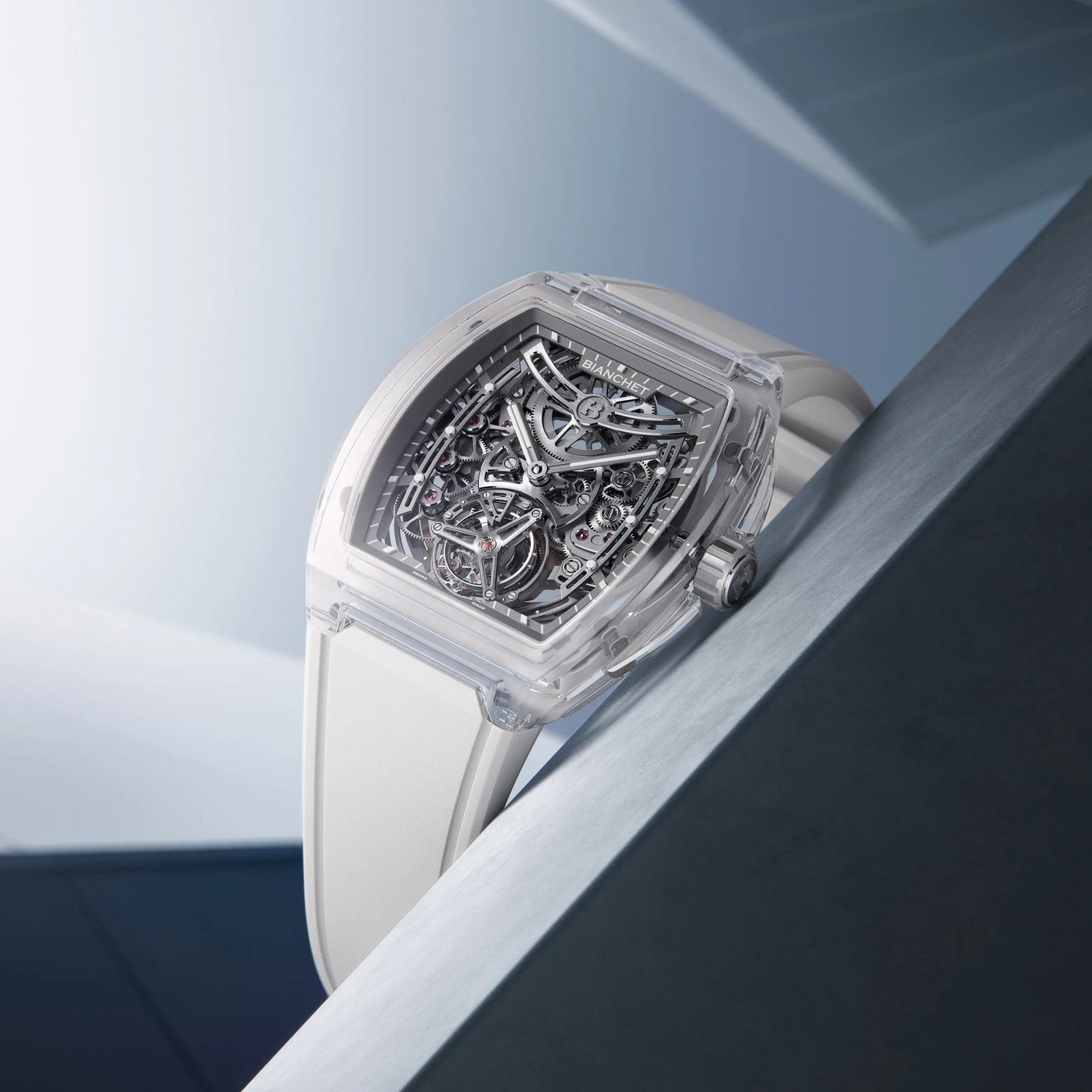 Transparent wristwatch with a white strap, featuring a visible intricate mechanical movement inside a rectangular case. The dial showcases gears and components with a modern, technical design, and the brand name "Bianchet" is visible near the top. The watch is positioned at an angle against a sleek, metallic surface with a soft gradient background.
