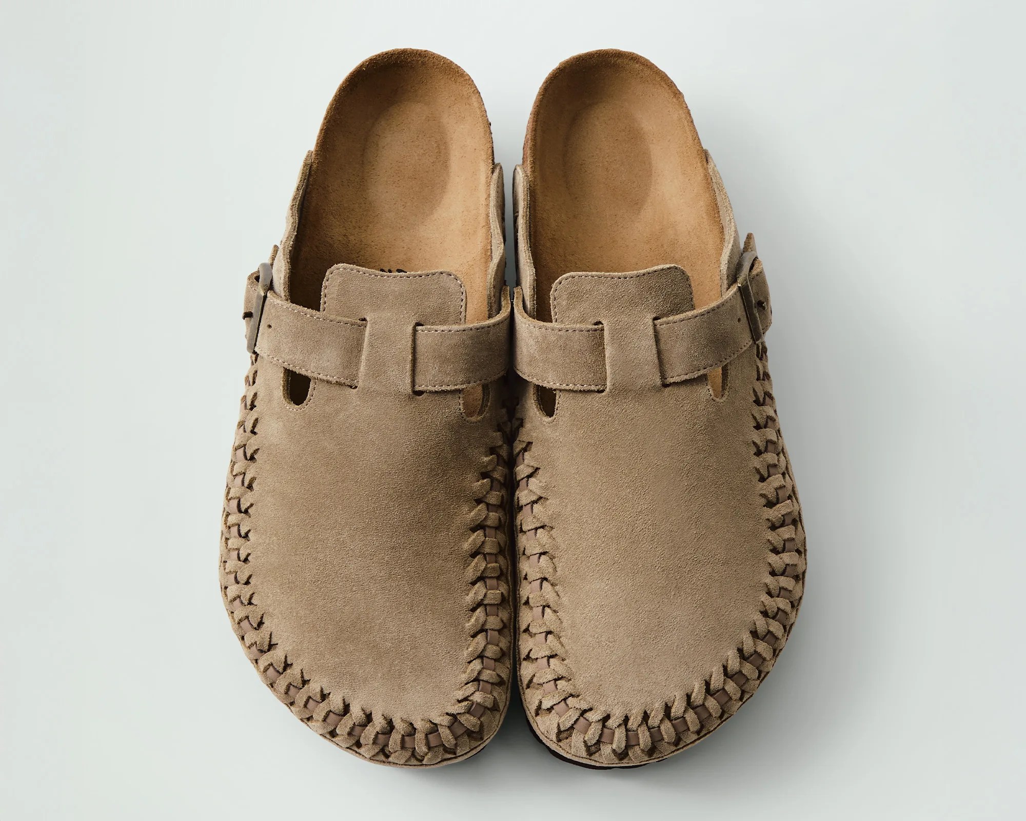 Pair of light brown suede slip-on shoes with woven detailing around the edges and a strap with a buckle across the top.