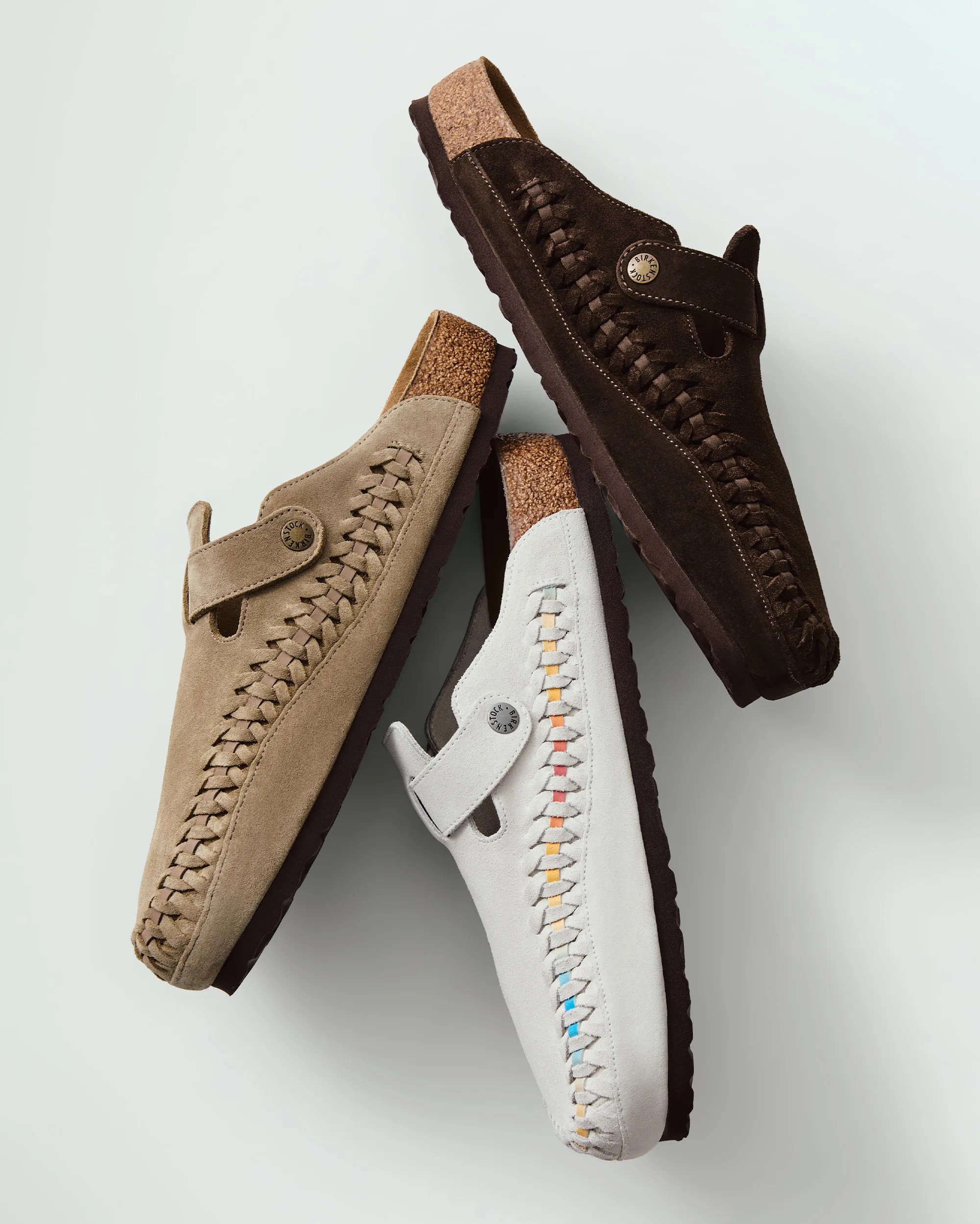 Three pairs of slip-on shoes with woven side details and cork footbeds are displayed. The shoes come in three colors: dark brown, beige, and light gray with multicolored accents woven into the side. Each shoe has a strap with a round metal button featuring the brand name "Birkenstock." The soles are dark brown with a textured grip.