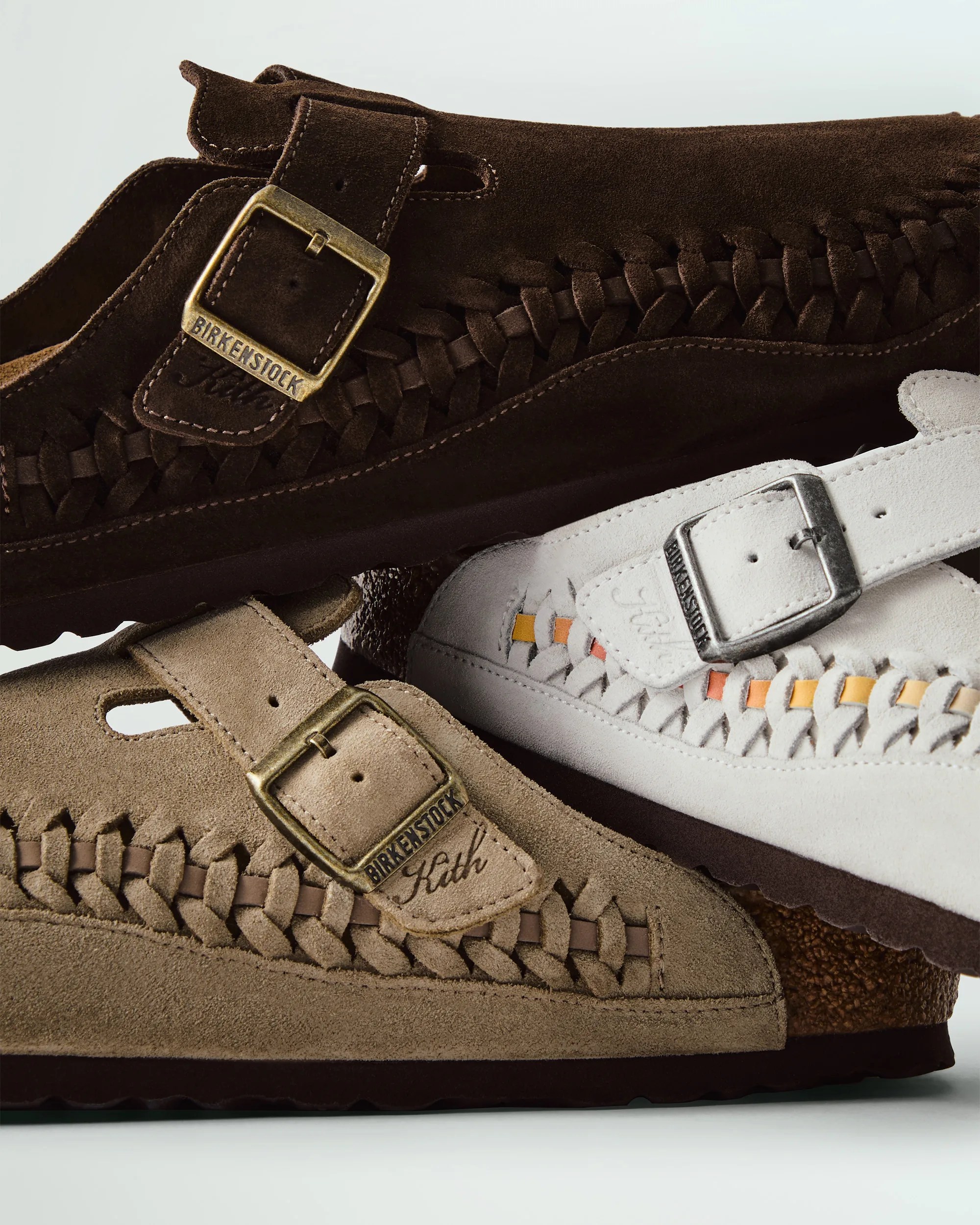 Three Birkenstock suede sandals in different colors—dark brown, beige, and white—are stacked together. Each sandal features a woven leather design along the side and a metal buckle with the Birkenstock logo. The beige sandal has "Kith" embroidered near the buckle. The white sandal includes small orange and yellow accents woven into the leather.