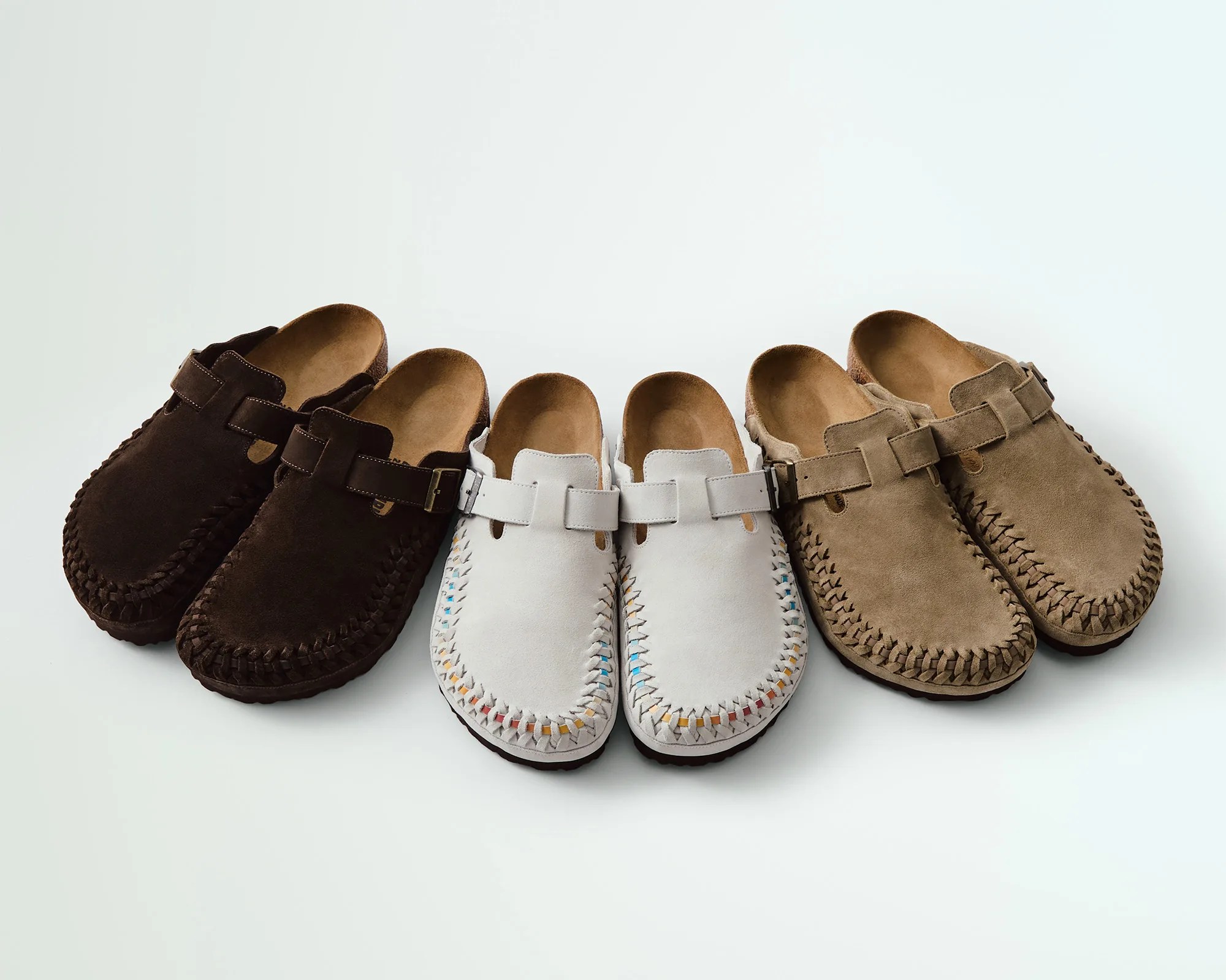 Three pairs of slip-on moccasin-style shoes with buckled straps are arranged side by side. The pair on the left is dark brown, the middle pair is light gray with colorful stitching along the edges, and the pair on the right is tan with matching stitching. All have a woven detail around the toe area and a soft suede texture.