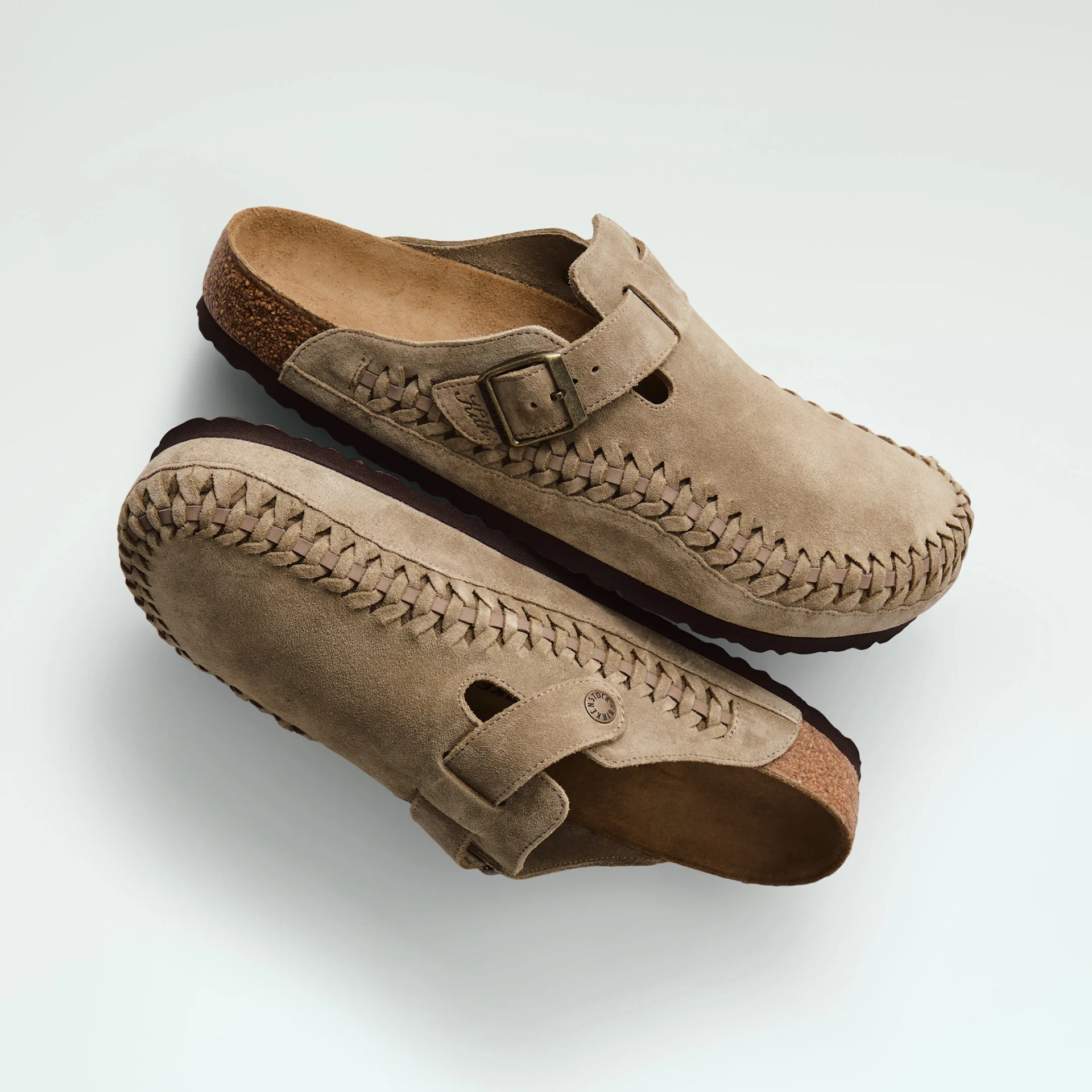 Pair of light brown suede slip-on clogs with woven leather detailing along the edges, a buckle strap on the top, and cork footbeds with dark brown rubber soles.