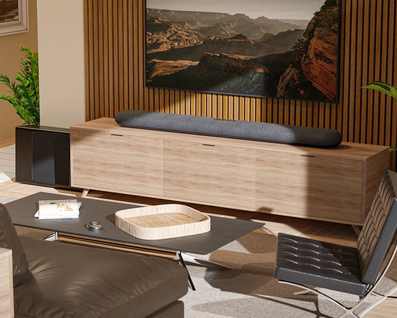 A modern living room setup featuring a wooden media console with a long, dark gray soundbar on top. A large flat-screen TV mounted on a vertical wooden slat wall displays a scenic canyon landscape. To the left of the console is a black subwoofer, and in front is a sleek black coffee table with a light wooden tray and a couple of books. The room includes a dark leather chair with a tufted design and a matching leather sofa partially visible in the foreground. Green plants add a touch of nature to the space.