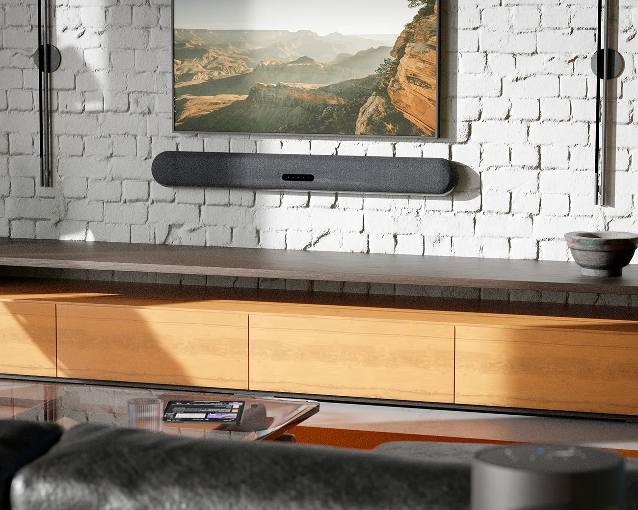 A modern living room setup with a wall-mounted flat-screen TV displaying a canyon landscape. Below the TV is a sleek, black soundbar attached to a white brick wall. A long wooden cabinet with a dark countertop runs beneath the soundbar. On the right side of the cabinet, there is a decorative bowl. The foreground shows part of a glass coffee table with a tablet and a glass on it, and a dark sofa.