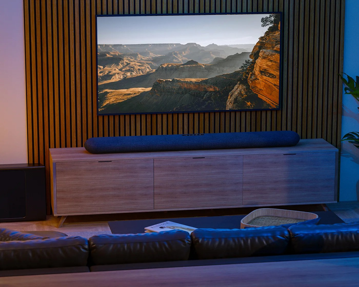 A modern living room setup featuring a large flat-screen TV mounted on a wooden slatted wall, displaying a scenic canyon landscape. Below the TV is a light wood media console with three cabinet doors, topped with a long, dark blue soundbar. In front of the console is a dark coffee table with a book and a woven tray. The edge of a dark leather sofa is visible in the foreground. The room has a cozy, ambient lighting with a mix of warm and cool tones.