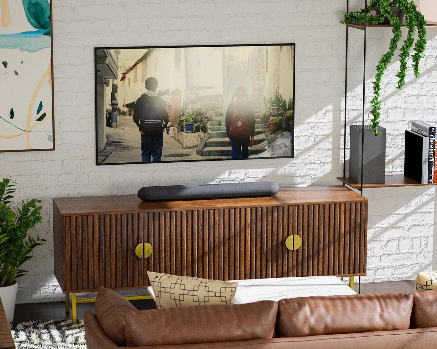 Wooden TV console with vertical slats and round brass handles, supported by brass legs. A black soundbar sits on top. Mounted above is a flat-screen TV displaying an image of two people walking up stairs. To the right, a glass shelf holds a black speaker, books, and a hanging green plant. A brown leather sofa with patterned cushions is in the foreground, and a potted plant is on the left. The background features a white brick wall with a framed abstract painting partially visible on the left.