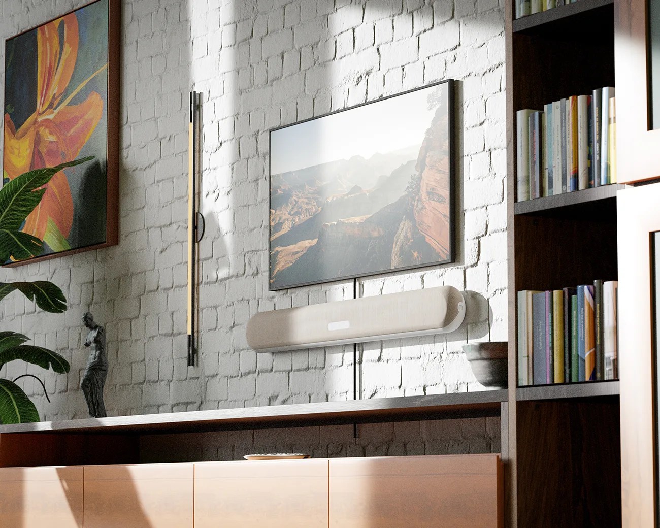 A modern living room wall features a mounted flat-screen TV displaying a scenic canyon landscape. Below the TV, a beige soundbar is mounted on the white brick wall. To the left, there is a vertical wall light fixture and a large framed painting of an orange flower with green leaves. A leafy green plant and a small statue are placed on a wooden cabinet beneath the TV. On the right side, a dark wooden bookshelf filled with books is partially visible.
