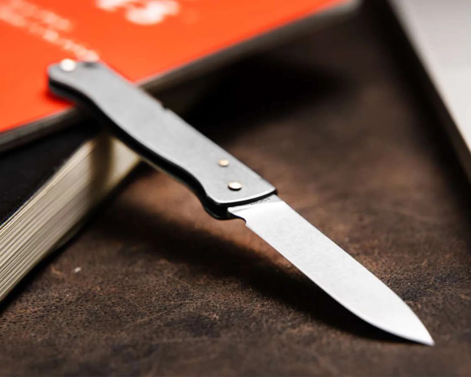 A folding knife with a silver blade and a black handle resting on a brown textured surface next to a book with a red cover.