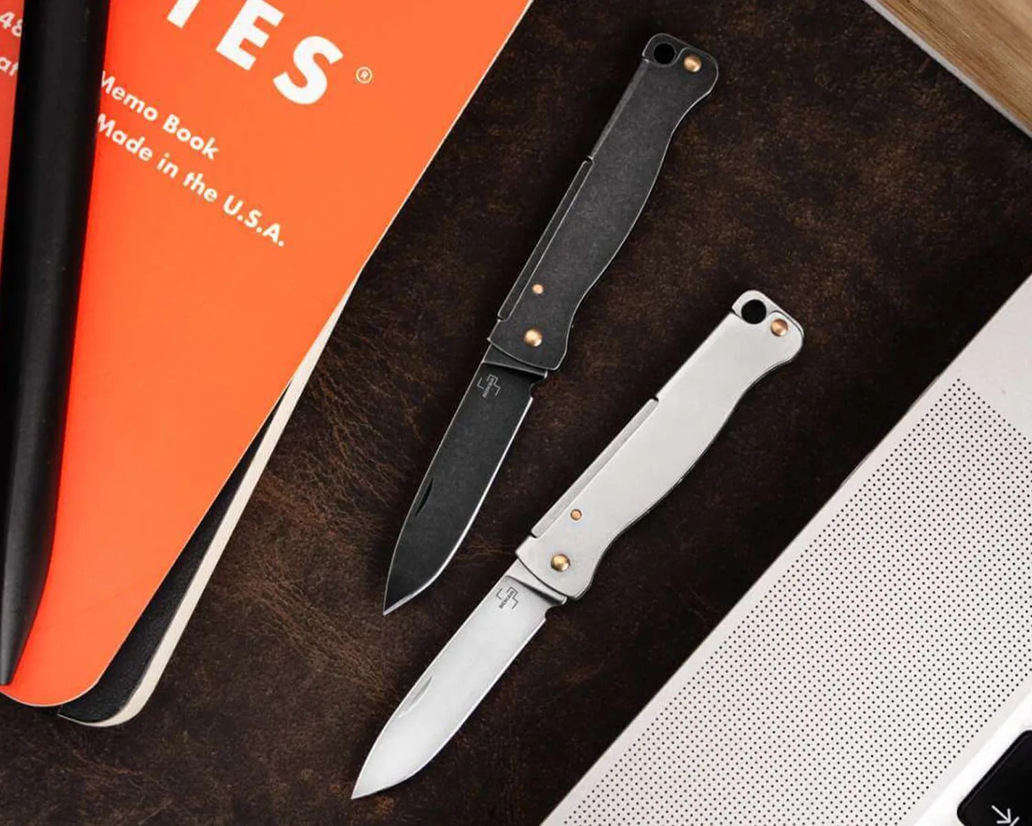 Two folding knives are placed on a dark brown leather surface. One knife has a black handle and blade, while the other has a silver handle and blade. Both knives have a small circular hole near the top of the handle and brass-colored rivets. To the left, there is an orange notebook partially visible with white text that reads "Memo Book Made in the U.S.A." and a black pen resting on it. On the right side, part of a silver laptop with a perforated speaker grill is visible.