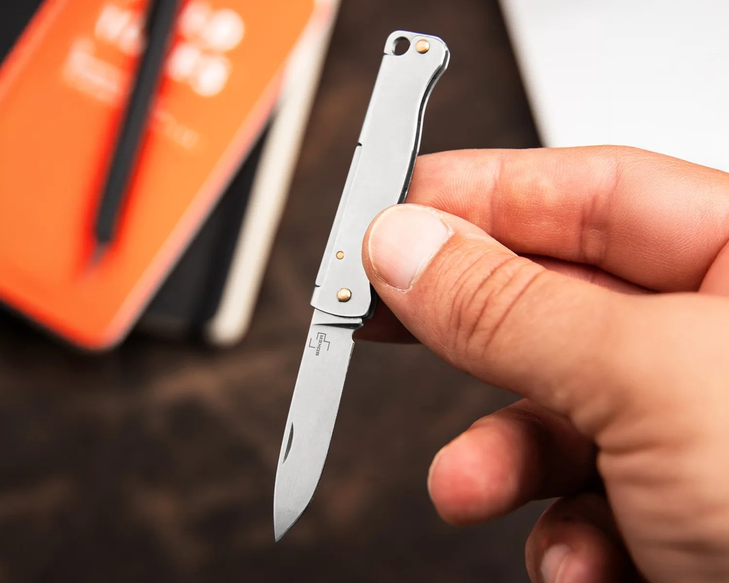 A hand holding a small folding pocket knife with a silver handle and a partially open blade. The blade has a logo and the word "Böker" on it. The background is blurred, showing some orange and black objects.
