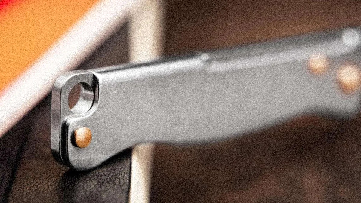 Close-up of a metallic folding knife handle with a circular hole near the end and brass rivets, resting on a textured surface. The background is blurred with warm tones.