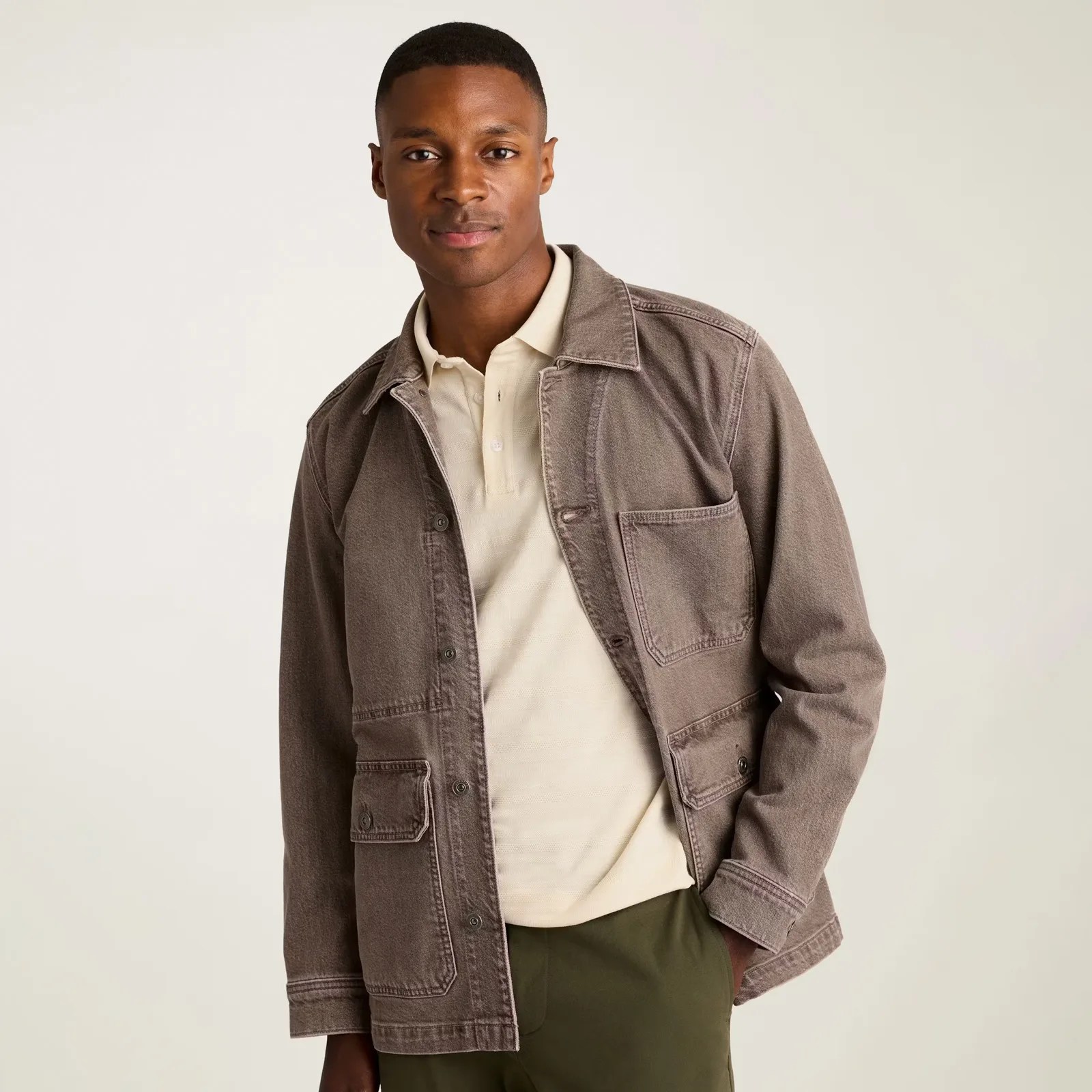 A man wearing a light beige polo shirt under a brownish-gray denim jacket with multiple pockets, paired with olive green pants. The jacket has a collar and button closures. The background is plain and light-colored.