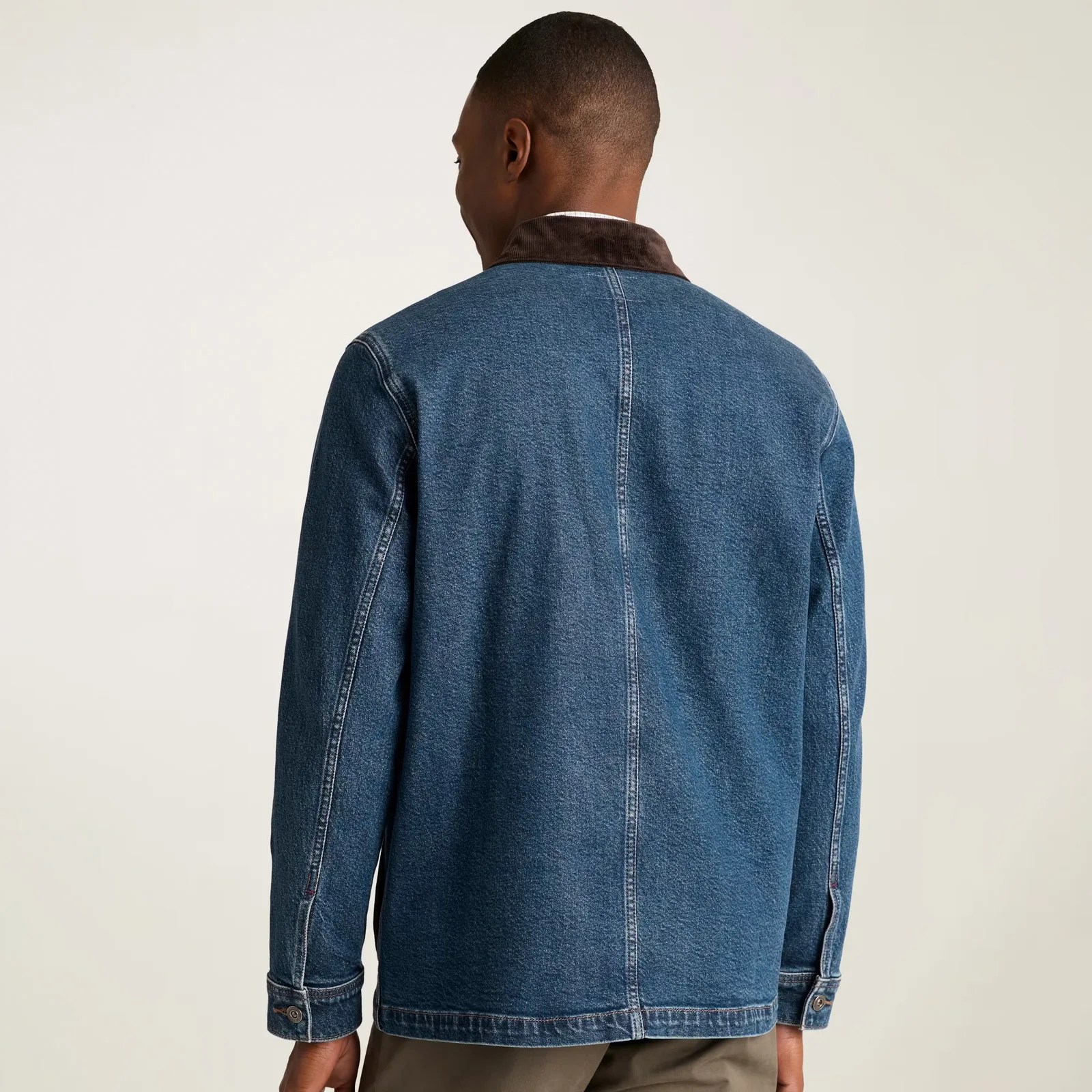 A man wearing a blue denim jacket with a dark brown corduroy collar, shown from the back. The jacket has visible stitching along the seams and cuffs with button closures. The man is also wearing light brown pants.