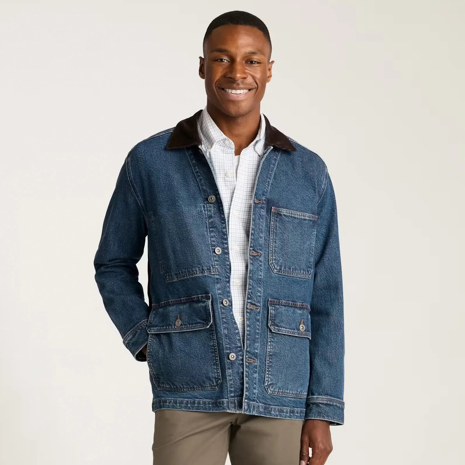 A man wearing a blue denim jacket with a dark brown collar over a white checkered button-up shirt. The jacket features multiple pockets, including two buttoned flap pockets at the bottom and one chest pocket. He is also wearing light brown pants.