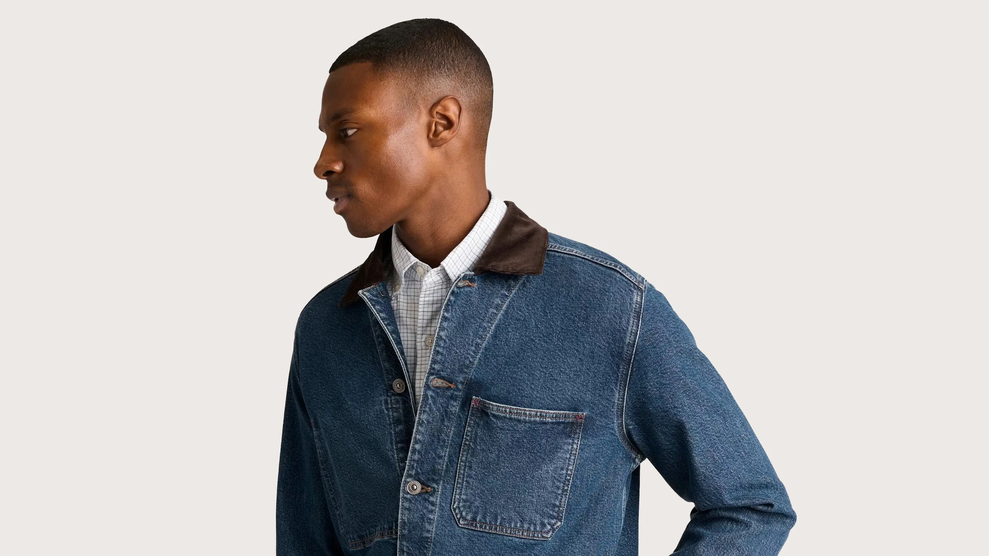 A man wearing a blue denim jacket with a dark brown collar over a white checkered button-up shirt, looking to his left.