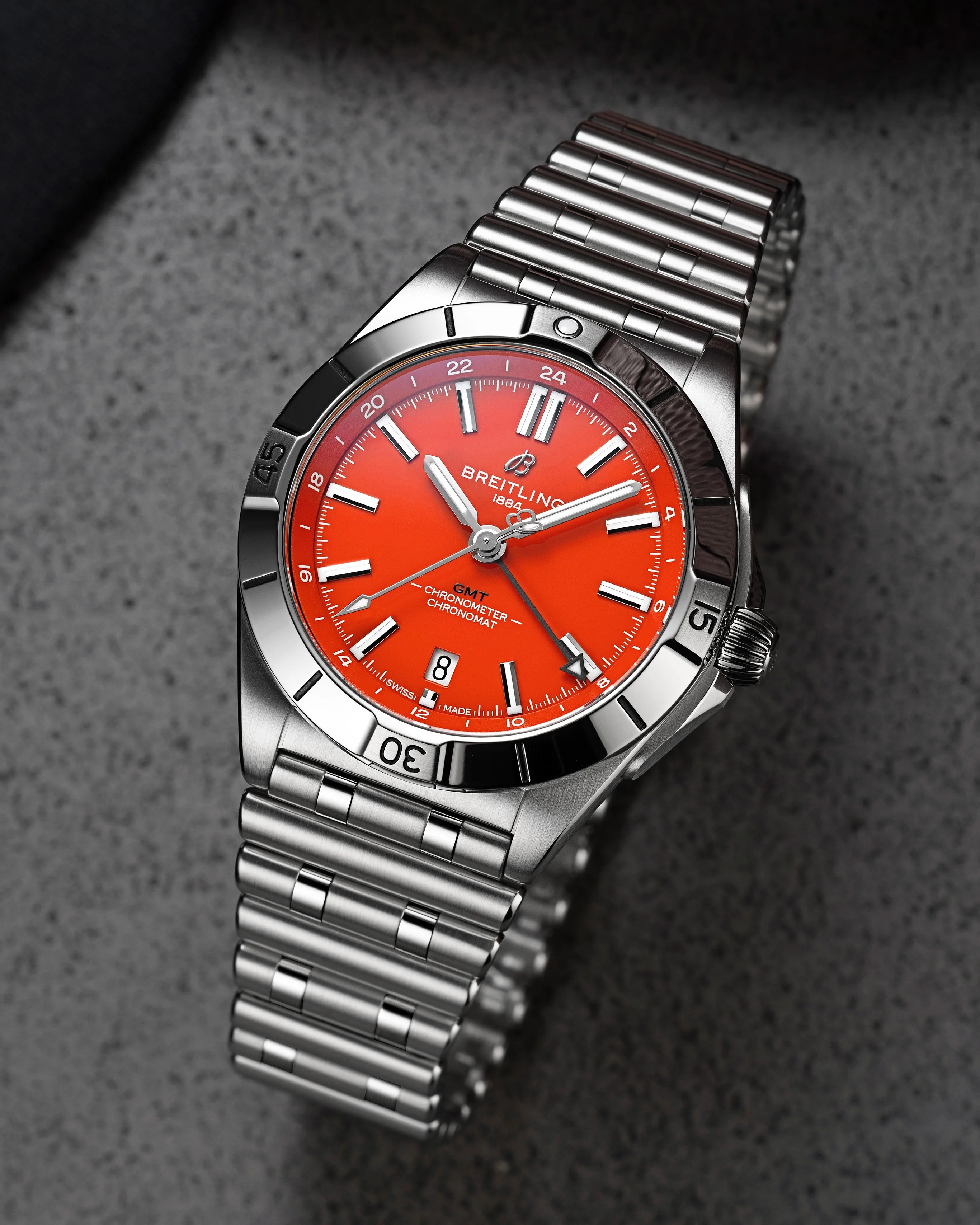 Breitling Chronomat GMT watch with a bright orange dial, silver hour markers, and hands. The watch features a stainless steel case and bracelet, a date window at 6 o'clock, and a rotating bezel with minute markers. The dial includes the Breitling logo, "1884," "GMT," "Chronometer," and "Chronomat" text.