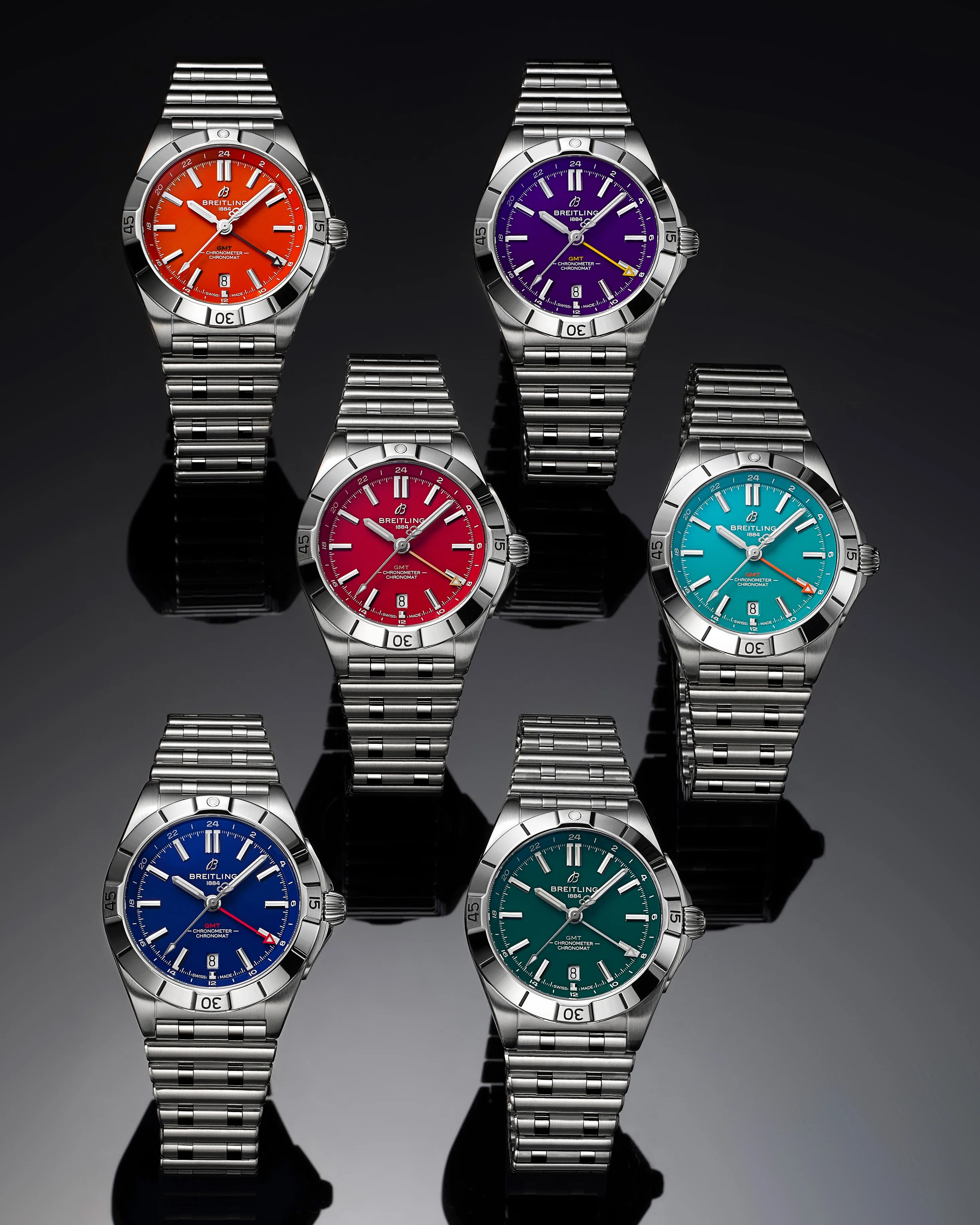 Six Breitling Chronomat GMT watches with stainless steel bracelets are displayed on a reflective black surface. Each watch features a different vibrant dial color: orange, purple, red, turquoise, blue, and green. All watches have silver hour markers and hands, a date window at 6 o'clock, and a rotating bezel. The GMT hand is highlighted in contrasting colors on each dial.