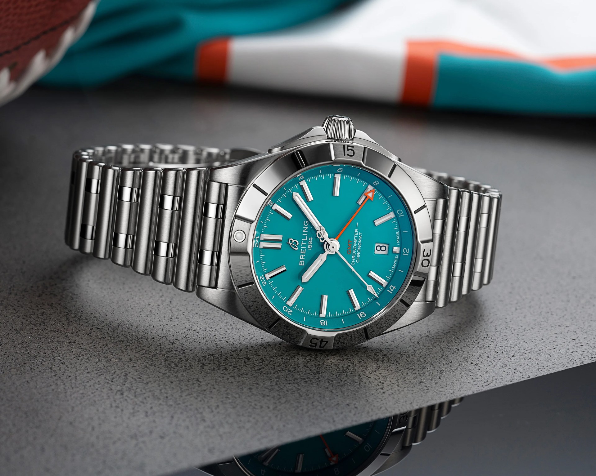 Stainless steel Breitling wristwatch with a turquoise dial, silver hour markers, and hands. The watch features a date window at 6 o'clock, an orange-tipped GMT hand, and a rotating bezel with engraved numerals. The bracelet is a polished and brushed metal link design. The watch is placed on a textured gray surface with a blurred background.