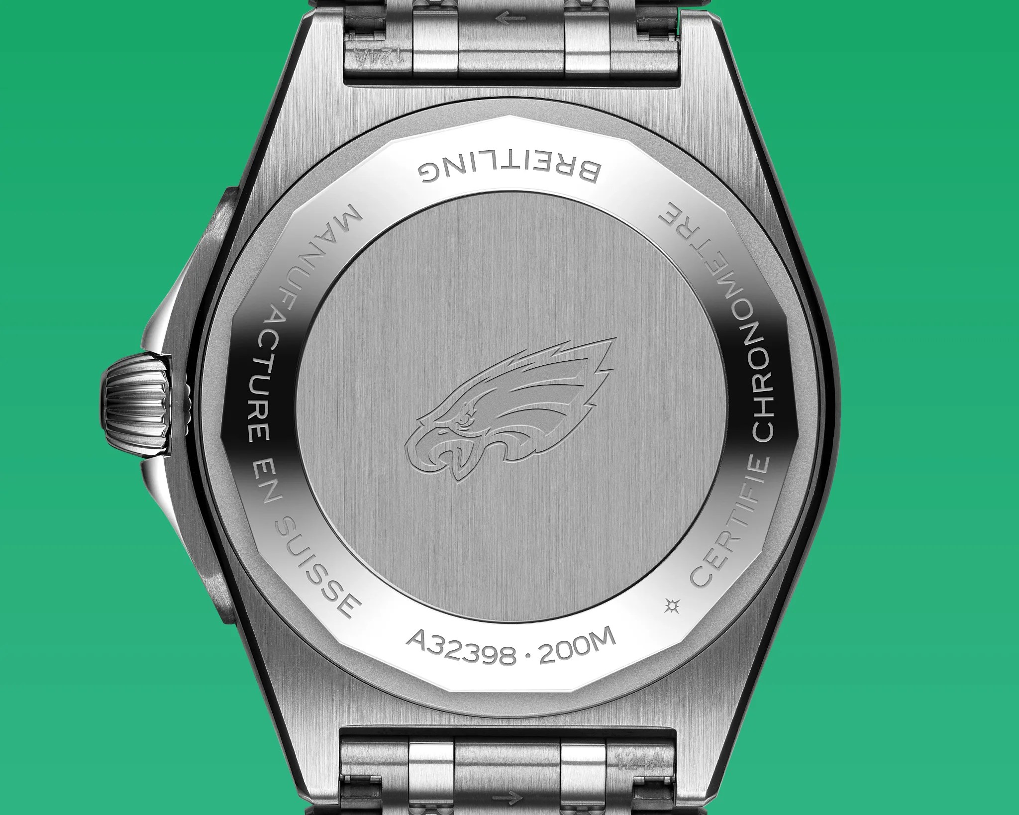 Stainless steel watch back with an engraved eagle head logo in the center. Around the edge, the text reads "BREITLING," "MANUFACTURE EN SUISSE," "A32398," "200M," and "CERTIFIE CHRONOMETRE." The watch has a metal bracelet and a crown on the left side. The background is green.