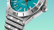 Close-up of a stainless steel wristwatch with a turquoise dial, featuring a date window at the 6 o'clock position, silver hour markers, and an orange GMT hand. The bezel is polished with black markings, and the watch has a brushed metal bracelet. The crown is ridged with an engraved logo. The text "GMT CHRONOMETER CHRONOMAT" is visible on the dial.