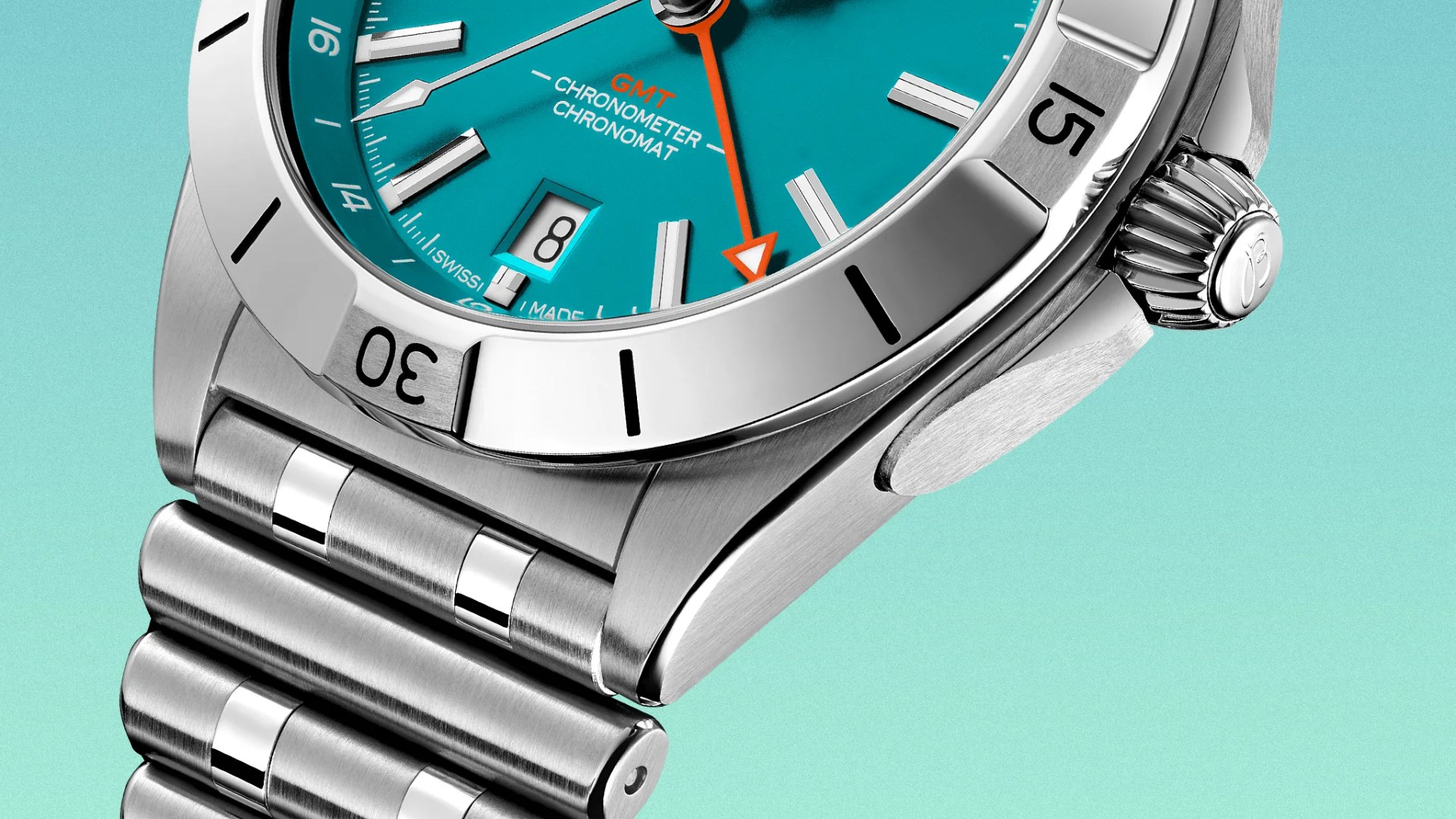 Close-up of a stainless steel wristwatch with a turquoise dial, featuring a date window at the 6 o'clock position, silver hour markers, and an orange GMT hand. The bezel is polished with black markings, and the watch has a brushed metal bracelet. The crown is ridged with an engraved logo. The text "GMT CHRONOMETER CHRONOMAT" is visible on the dial.