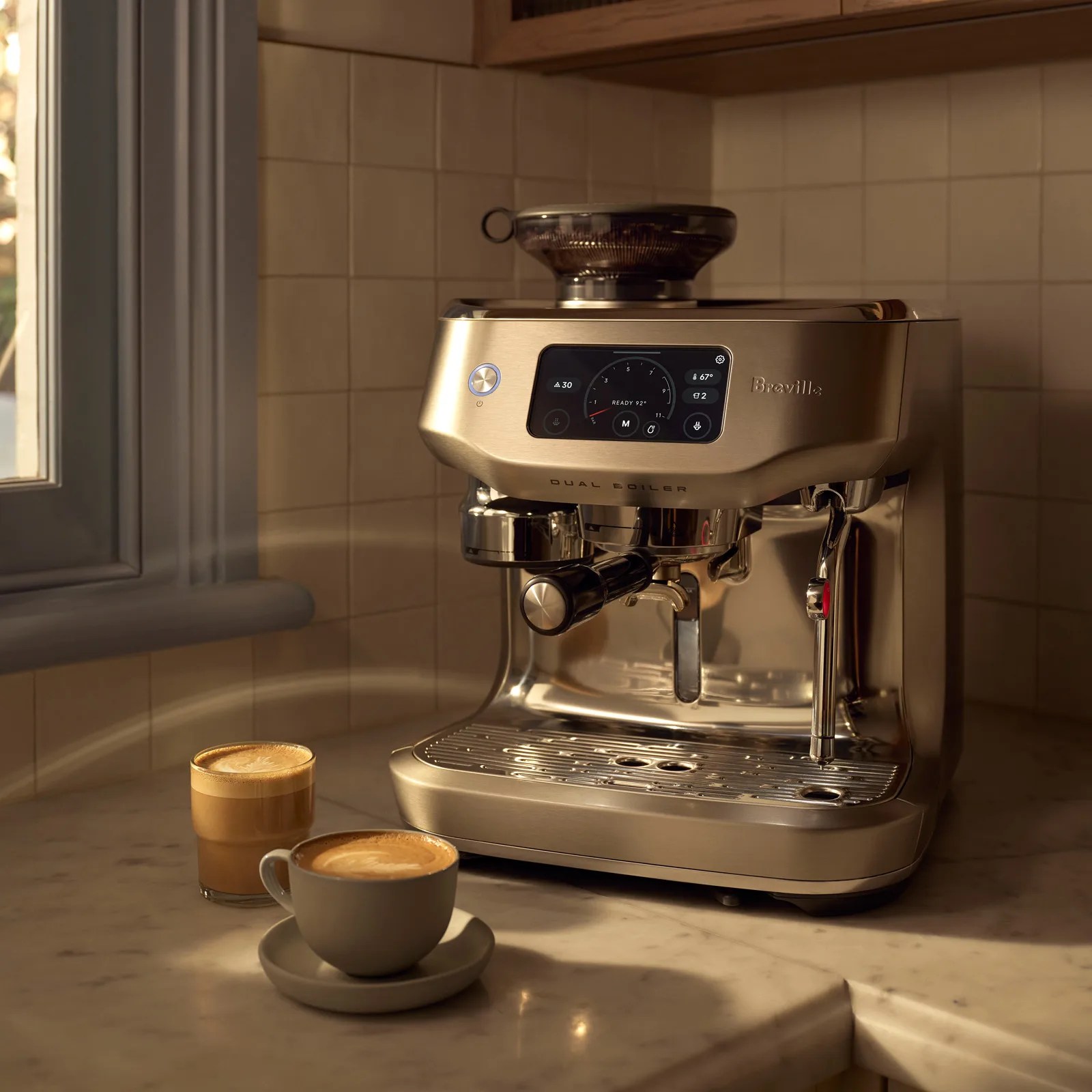 A sleek, metallic Breville espresso machine with a digital display and dual boiler system sits on a marble countertop in a kitchen corner. Next to it are two cups of coffee, one in a gray ceramic cup and saucer, and the other in a clear glass, both topped with latte art. The background features beige tiled walls and a window with gray trim.