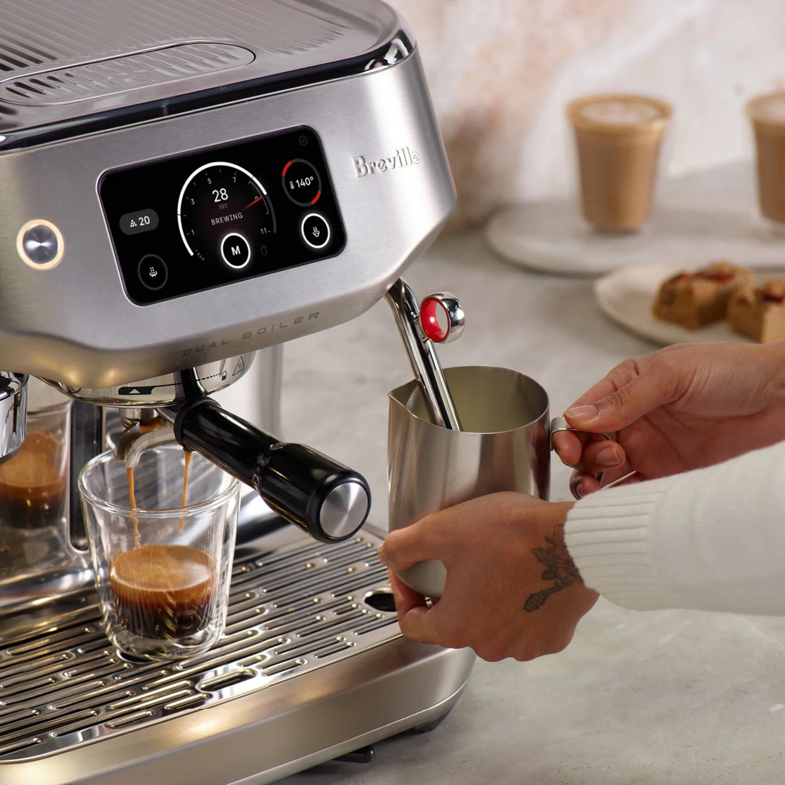 A stainless steel Breville espresso machine with a digital display showing brewing time and temperature. Espresso is being dispensed into a clear glass cup, while a person holds a metal milk frothing pitcher under the steam wand. The background includes two cups of coffee and a plate with pastries on a light-colored countertop.