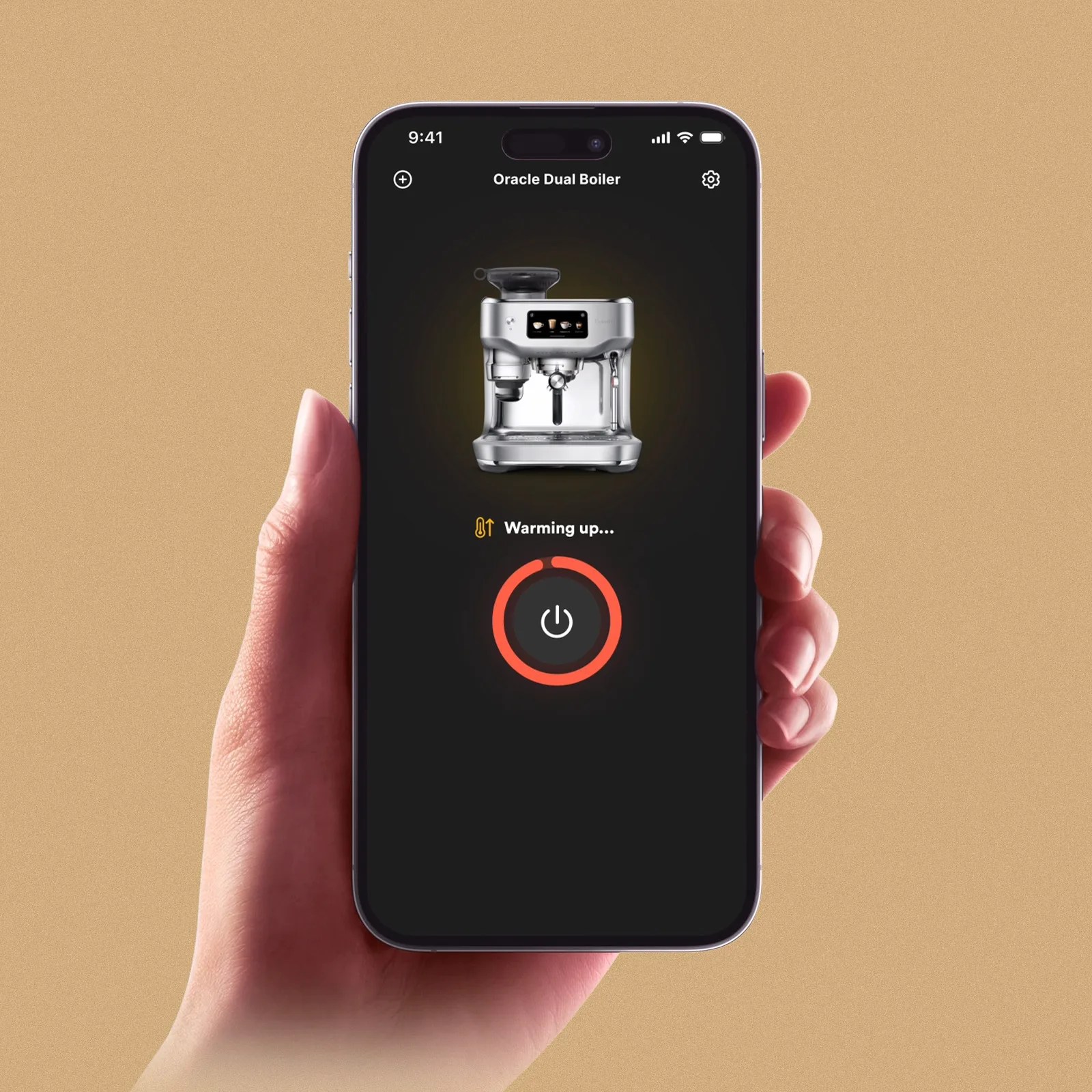 A hand holding a smartphone displaying an app interface for an "Oracle Dual Boiler" espresso machine. The screen shows an image of the espresso machine with a status message "Warming up..." and a circular progress indicator surrounding a power button icon. The background of the app is black with white and orange text and icons.