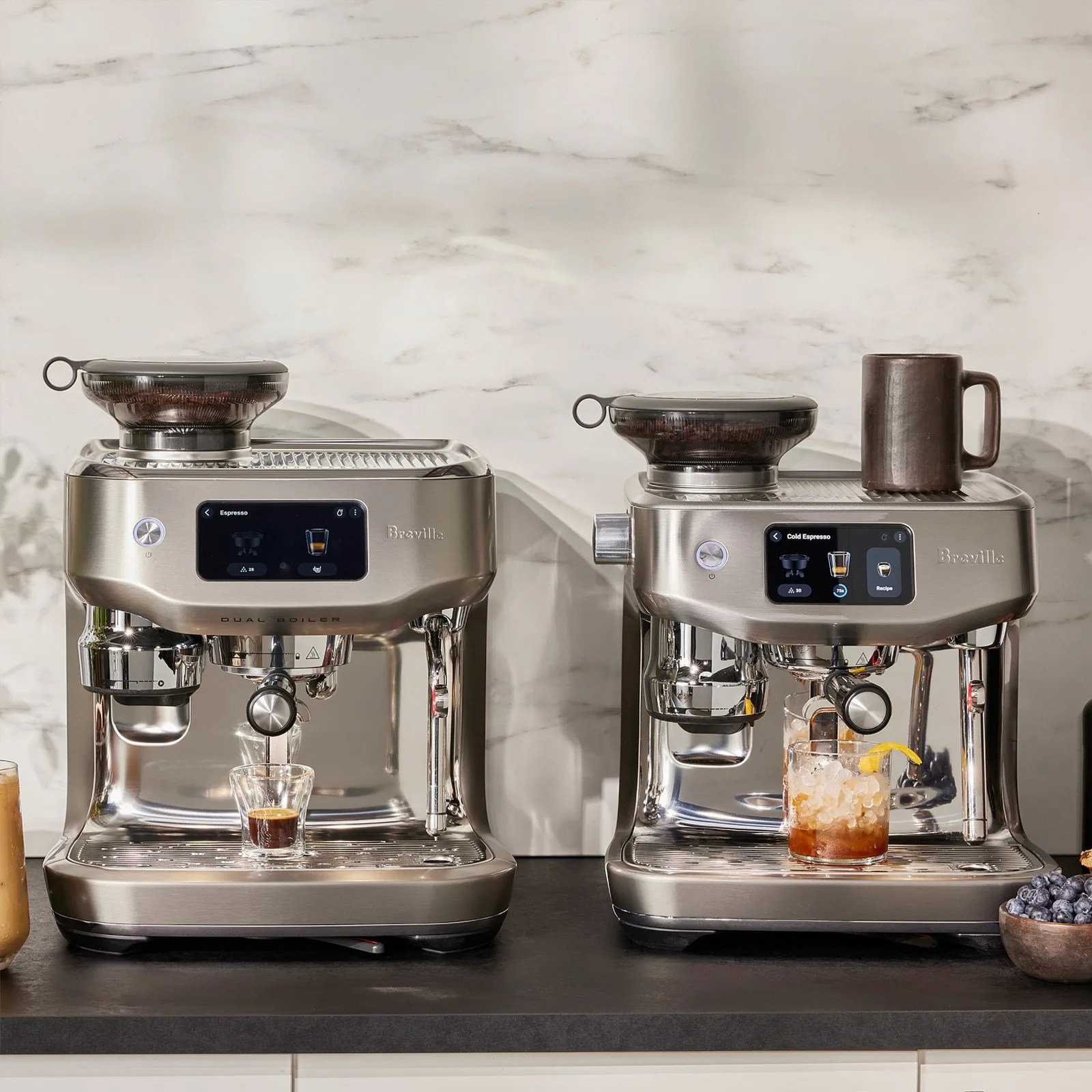 Two stainless steel Breville espresso machines with built-in grinders sit side by side on a dark countertop against a light marble backsplash. The machine on the left is brewing espresso into a small glass, while the machine on the right is dispensing a cold espresso drink into a glass filled with ice and garnished with a lemon twist. A brown ceramic mug rests on top of the right machine, and a small bowl of blueberries is visible on the far right of the counter.