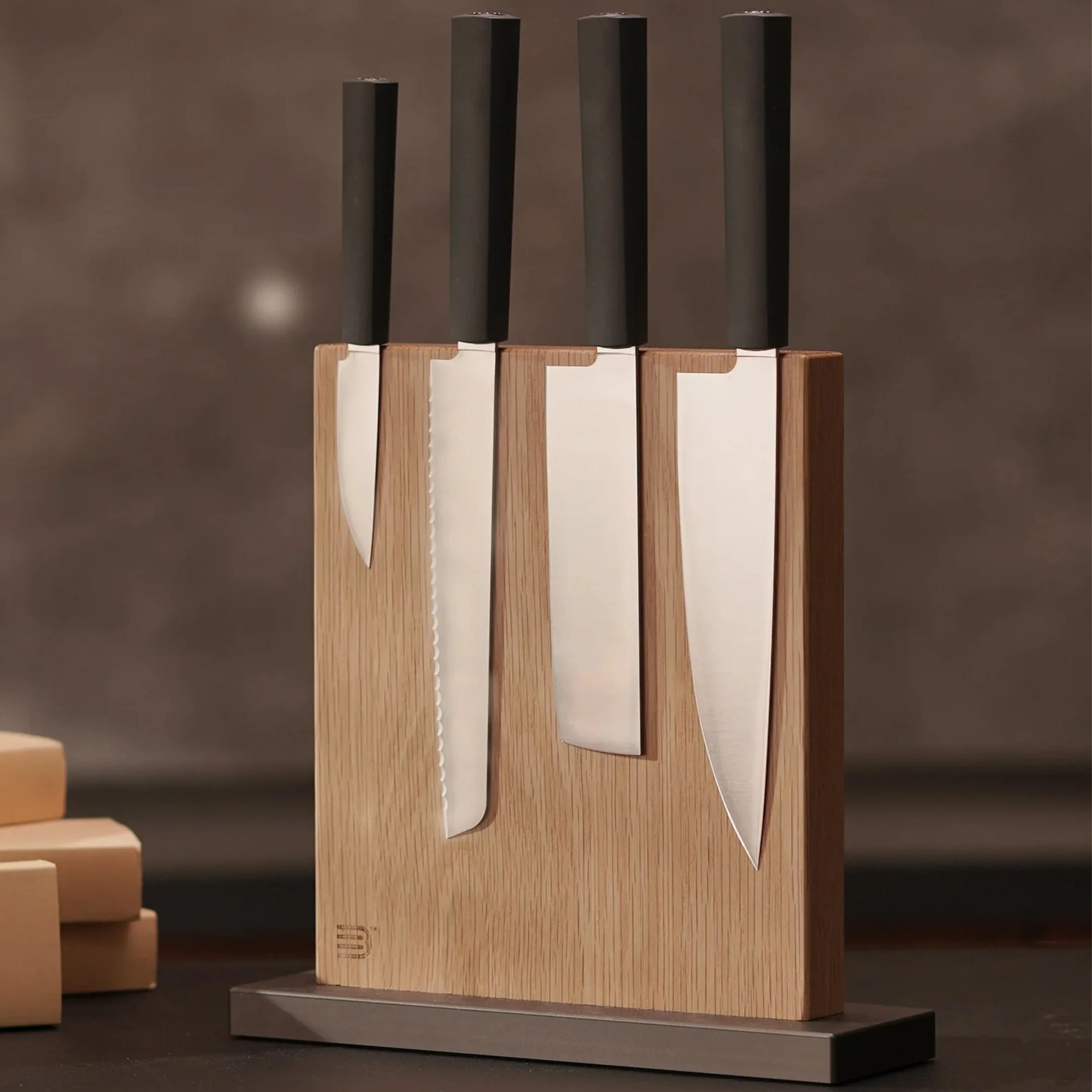Magnetic wooden knife block holding four kitchen knives with black handles and stainless steel blades, arranged vertically. The block has a natural wood finish and sits on a dark rectangular base.