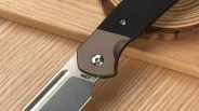Close-up of a folding knife showing the junction between the blade and the handle. The blade is metallic with a satin finish and has "CR3" engraved near the handle. The handle features a textured black grip and a smooth brown section secured with a silver screw. The knife rests on a wooden surface.