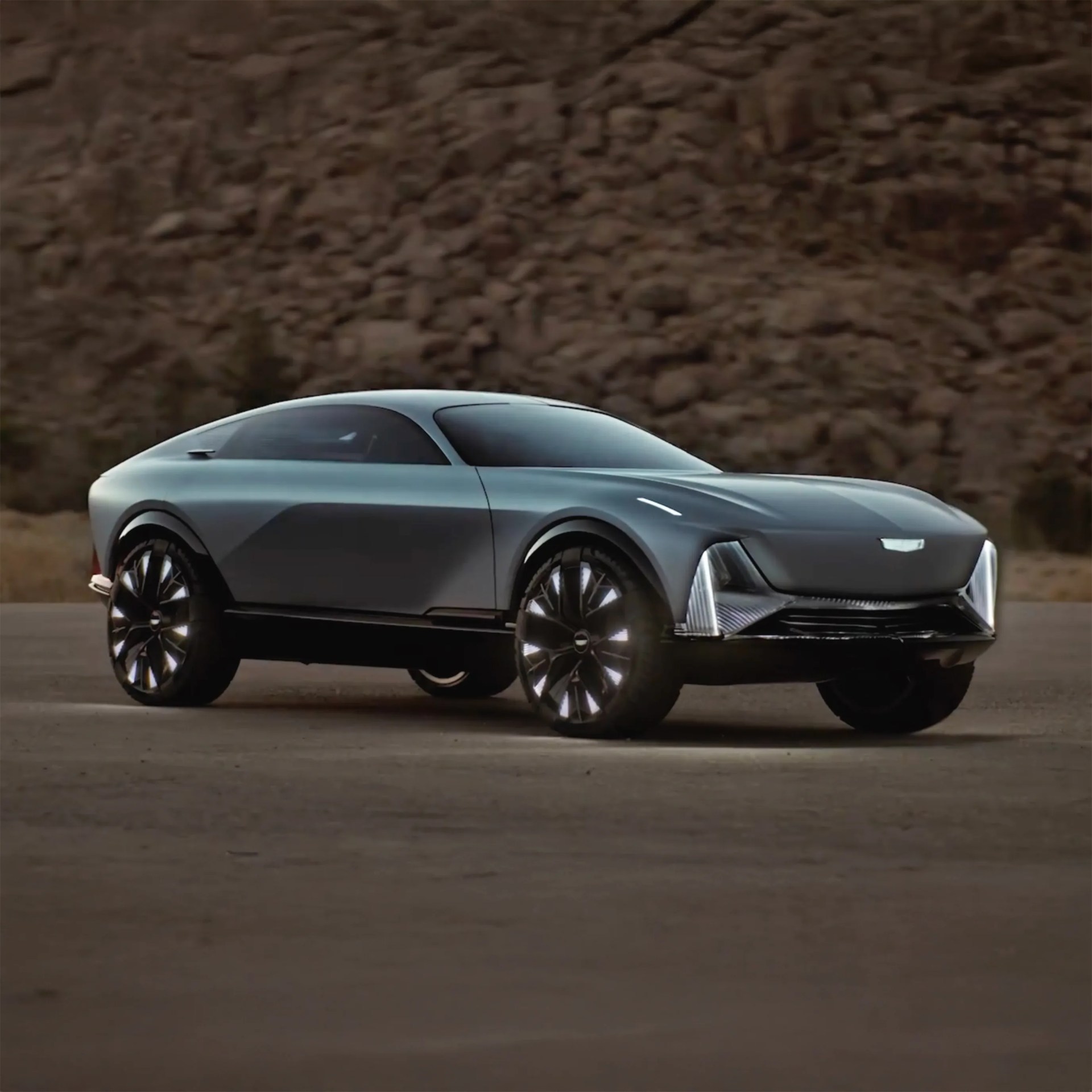 Sleek, futuristic electric SUV with a smooth, aerodynamic body in matte gray, large black alloy wheels with illuminated accents, and distinctive vertical LED headlights integrated into the front fascia. The vehicle has a coupe-like roofline and tinted windows, set against a rocky desert background.