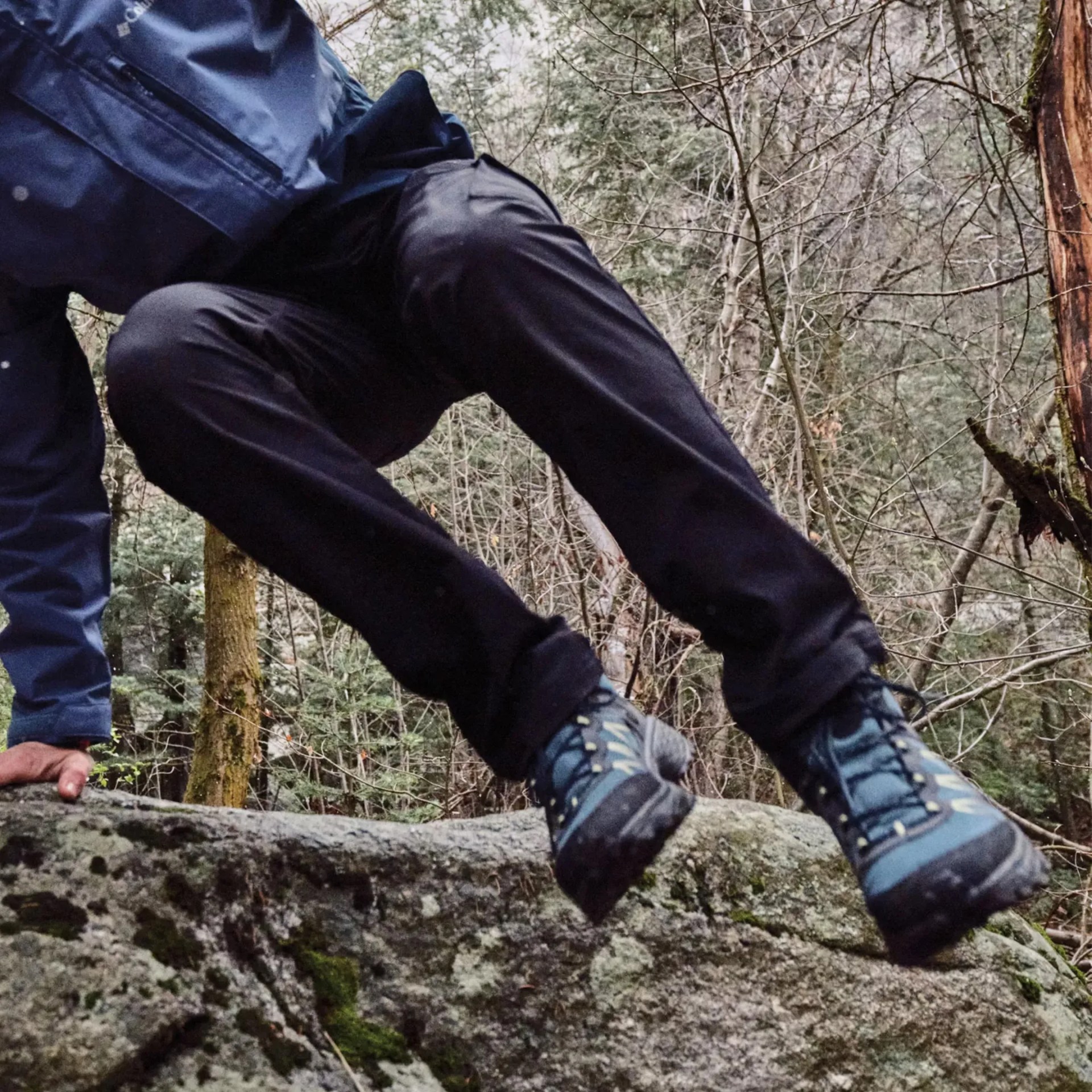 A person wearing black pants, blue hiking boots, and a blue jacket is climbing or jumping over a mossy rock in a forested area with bare trees and greenery in the background.