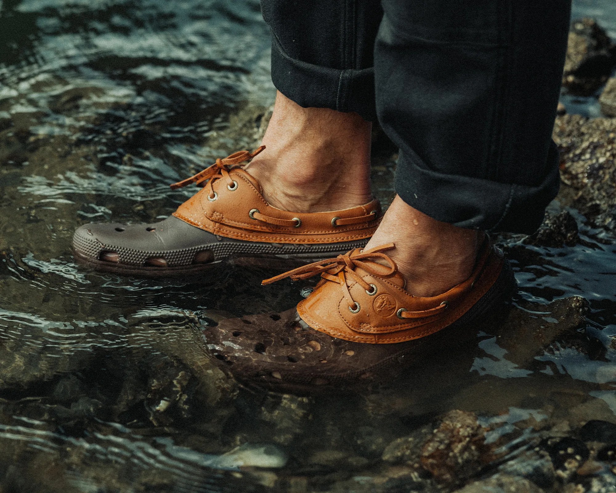 A pair of feet wearing brown and tan water-resistant boat shoes standing in shallow water with visible rocks underneath. The shoes have laces and ventilation holes, and the person is wearing black pants rolled up at the cuffs.