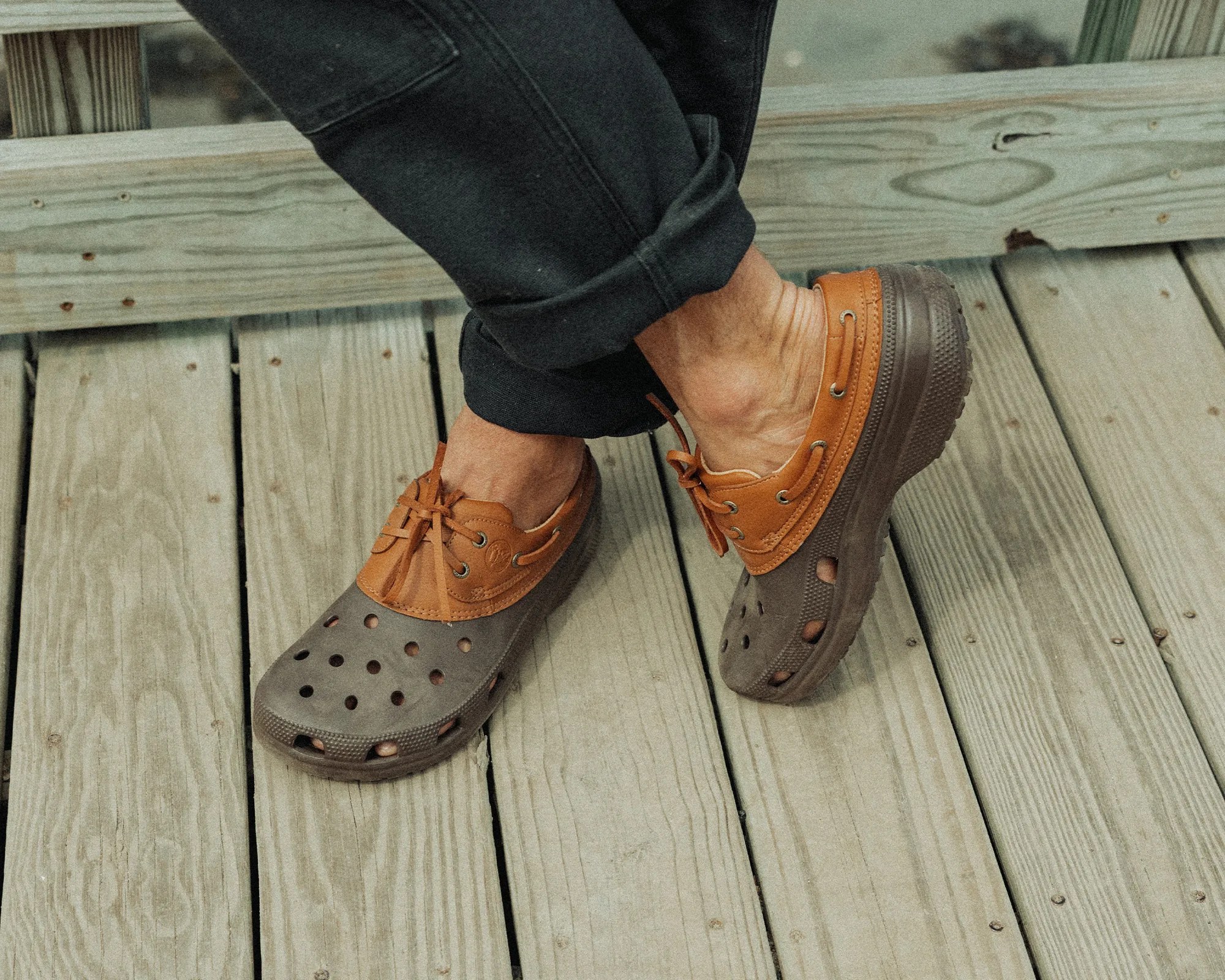 Brown and tan slip-on clogs with ventilation holes and lace-up details, worn with black rolled-up pants, on a wooden deck floor.