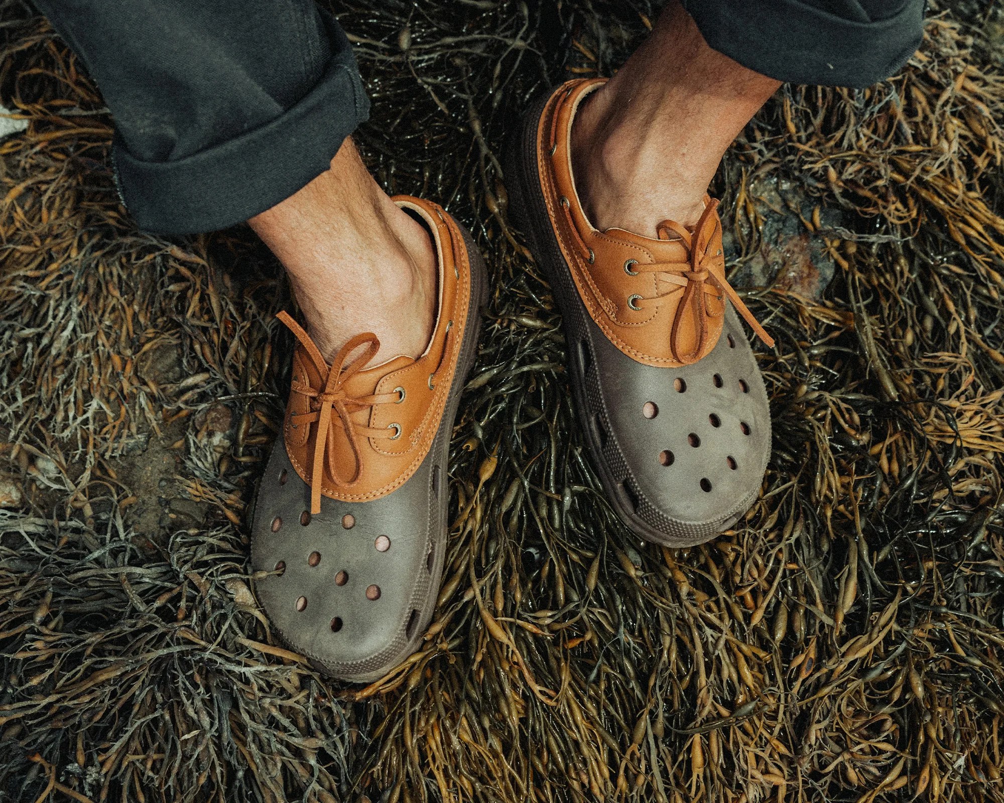 A pair of feet wearing two-tone slip-on shoes with brown leather laces and eyelets. The shoes have a gray base with multiple ventilation holes on the toe area. The person is standing on a bed of dark brown and green seaweed. The pants are rolled up slightly above the ankles.