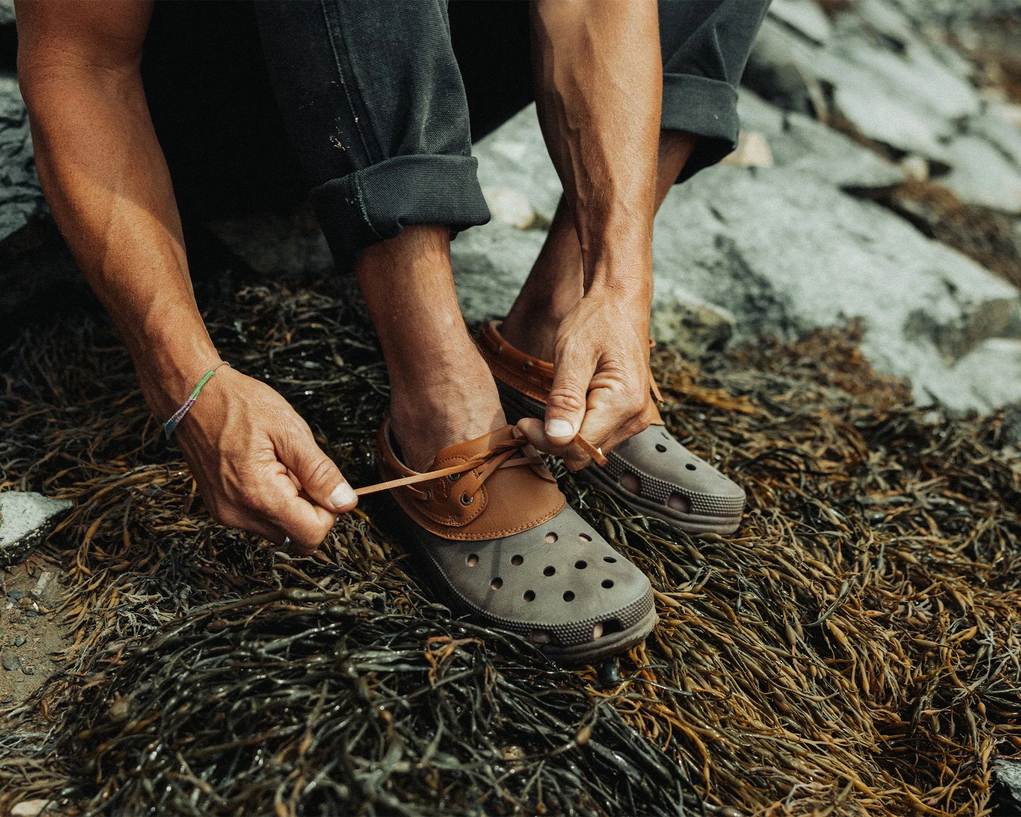 A person is tying the laces of brown and gray slip-on shoes with ventilation holes, sitting on a surface covered with seaweed and rocks. The person is wearing dark rolled-up pants and a multicolored woven bracelet on their left wrist.